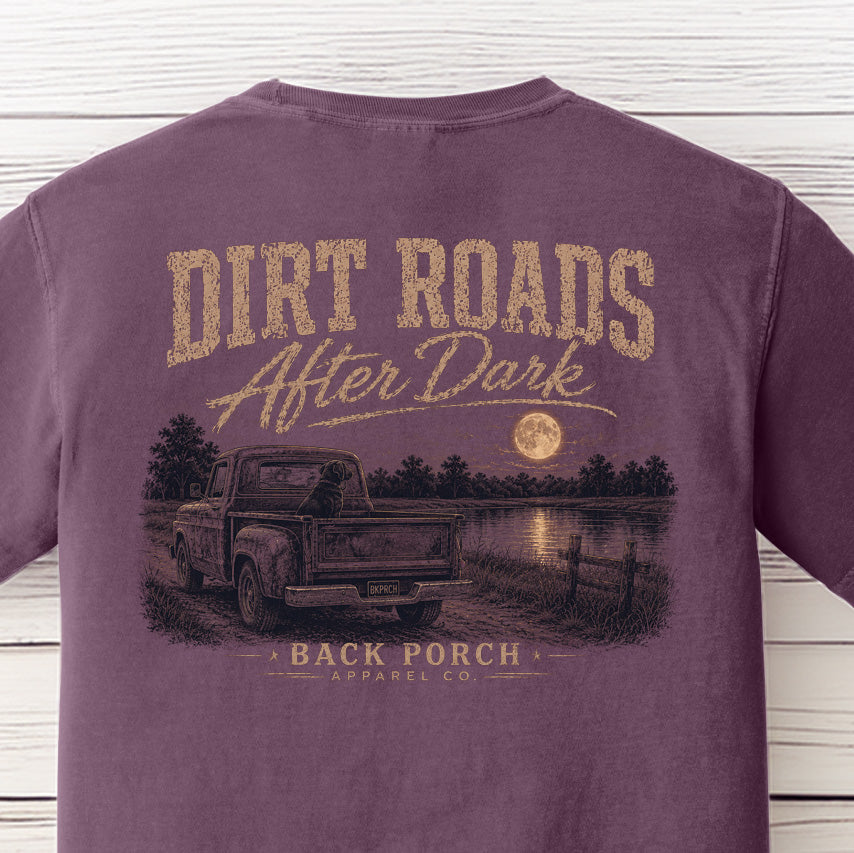 Close-up of the Dirt Roads After Dark graphic on a Berry Comfort Colors t-shirt showing an old pickup truck, black Labrador, moonlit lake, and Back Porch Apparel Co. branding.