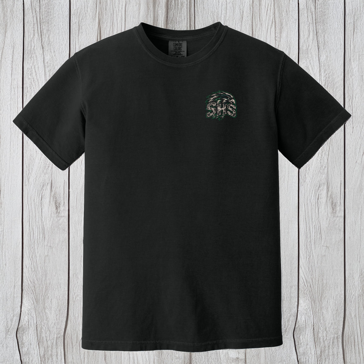 A black short-sleeve crewneck t-shirt with a small camouflage-patterned circular chest graphic, displayed flat against a light gray wood plank background.