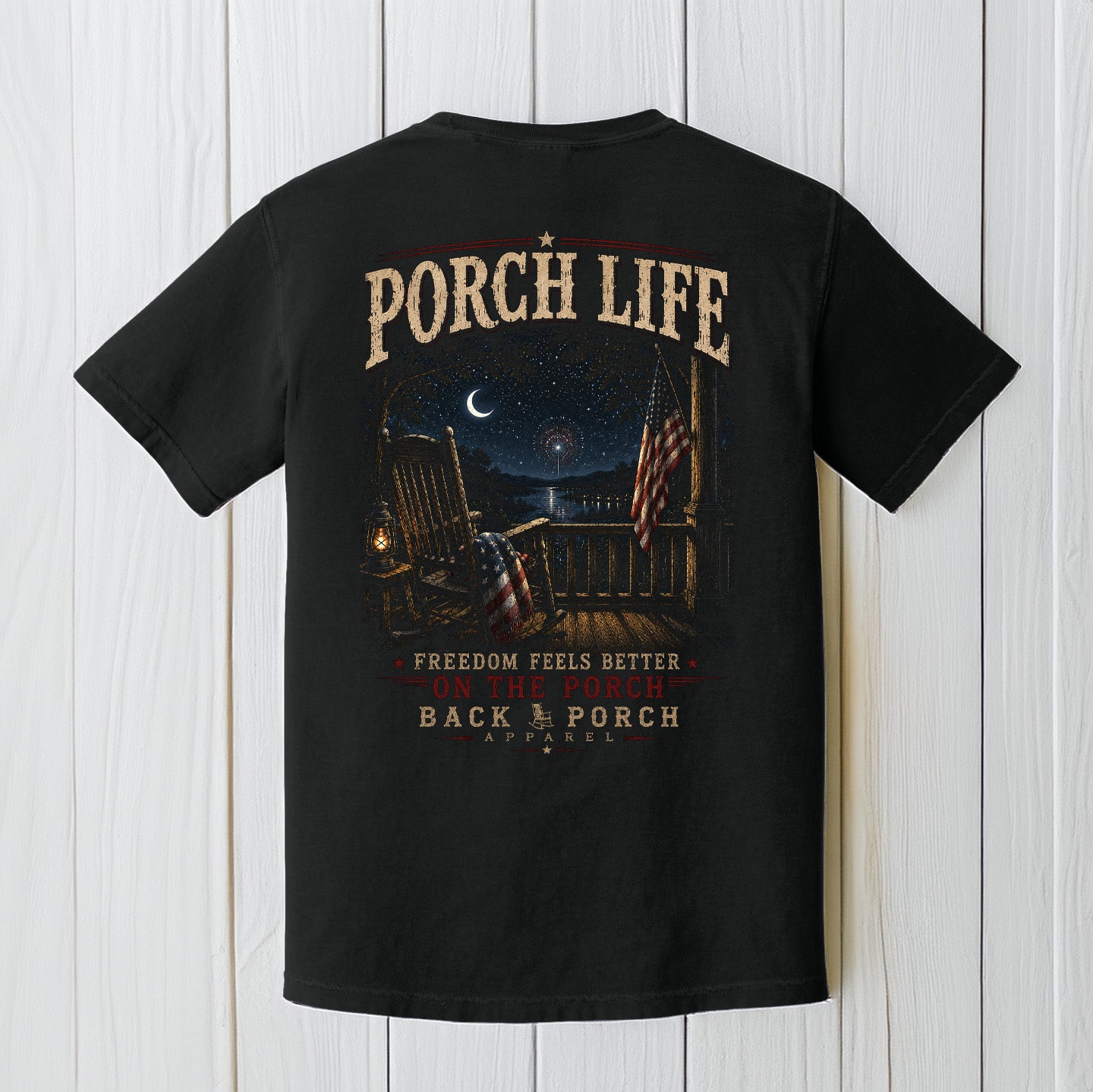 Black Comfort Colors t-shirt featuring patriotic porch life artwork displayed on a white wood background.