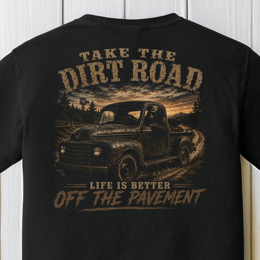 Close-up of the Take the Dirt Road graphic showing Bigfoot driving a vintage truck on a winding dirt road at sunset on a Black Comfort Colors t-shirt.