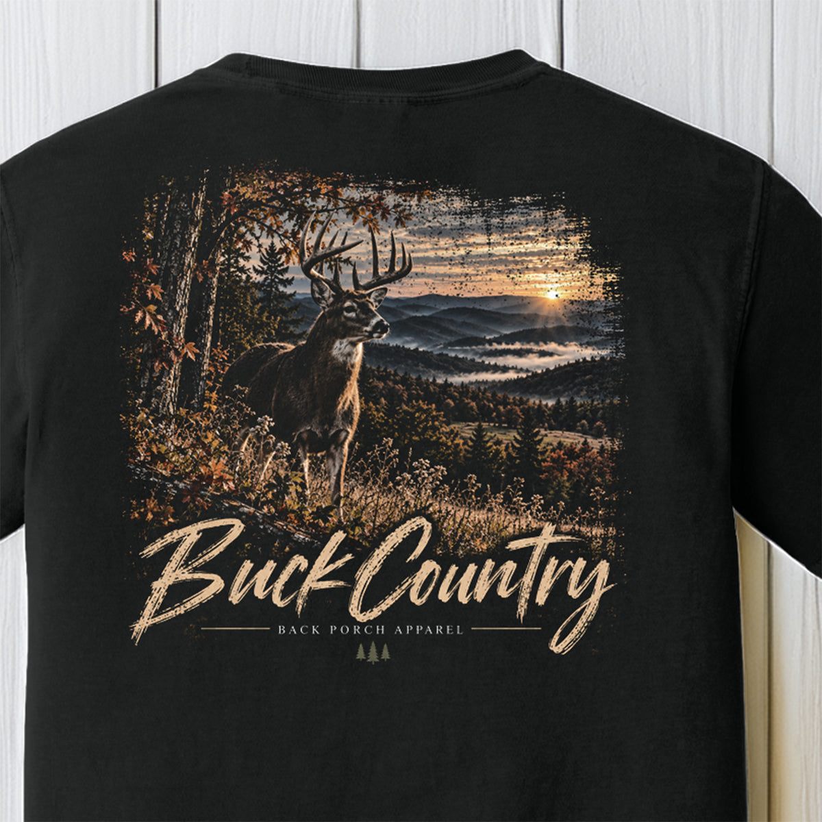 A black short-sleeve t-shirt featuring a large graphic print on the back. The graphic depicts a deer in a forest at sunset with the text 'Buck Country' and 'BACK PORCH APPAREL' underneath, displayed against a white wood-paneled background.
