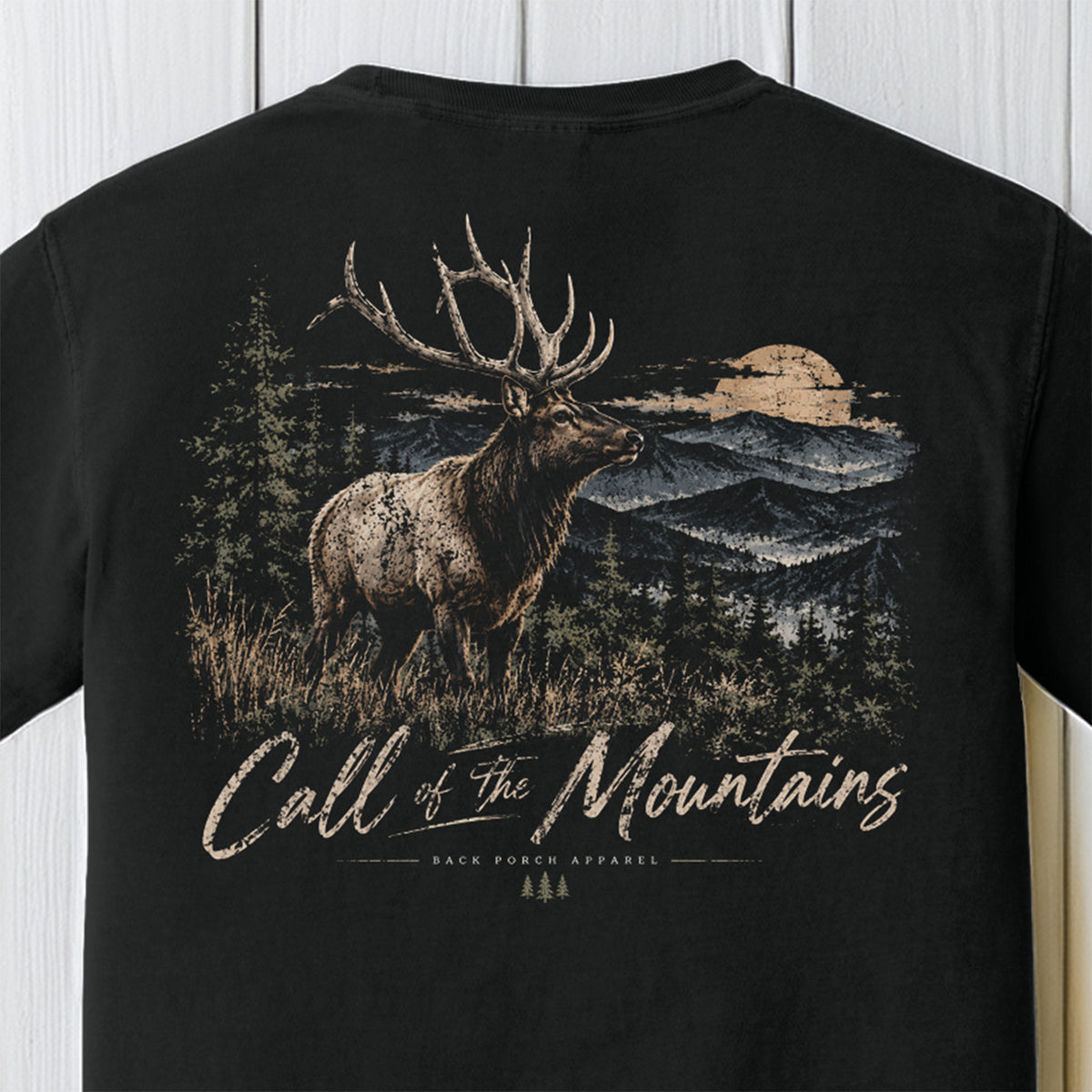 A black short-sleeve t-shirt featuring a large graphic print on the back. The graphic depicts a large elk with antlers in a mountain landscape at sunset, with the text 'Call of The Mountains' and 'BACK PORCH APPAREL' printed below.
