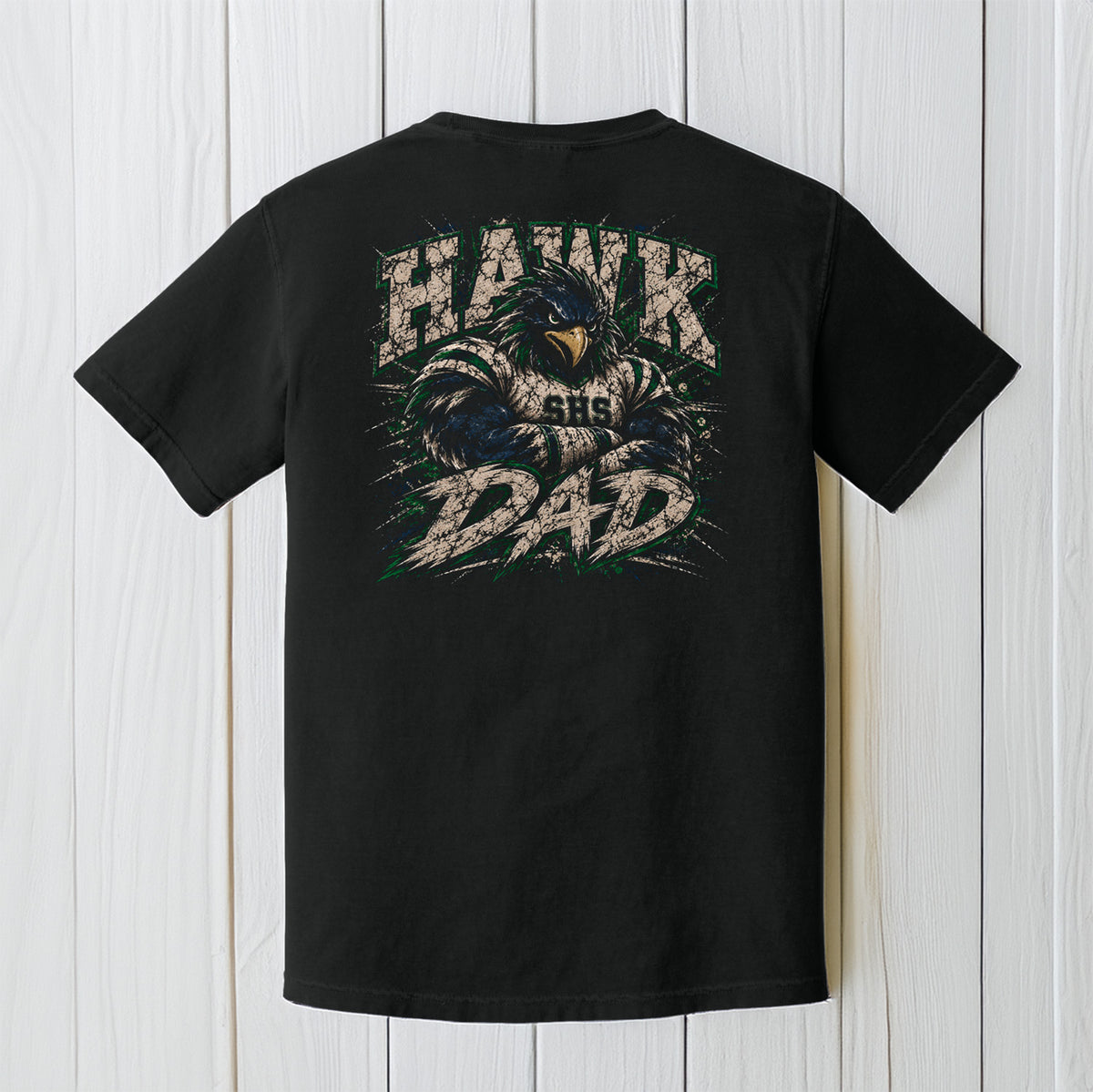 A black short-sleeve t-shirt shown from the back, featuring a large graphic print of a hawk with 'HAWK DAD' written in distressed white and green lettering. The t-shirt is displayed flat against a white wood plank background.