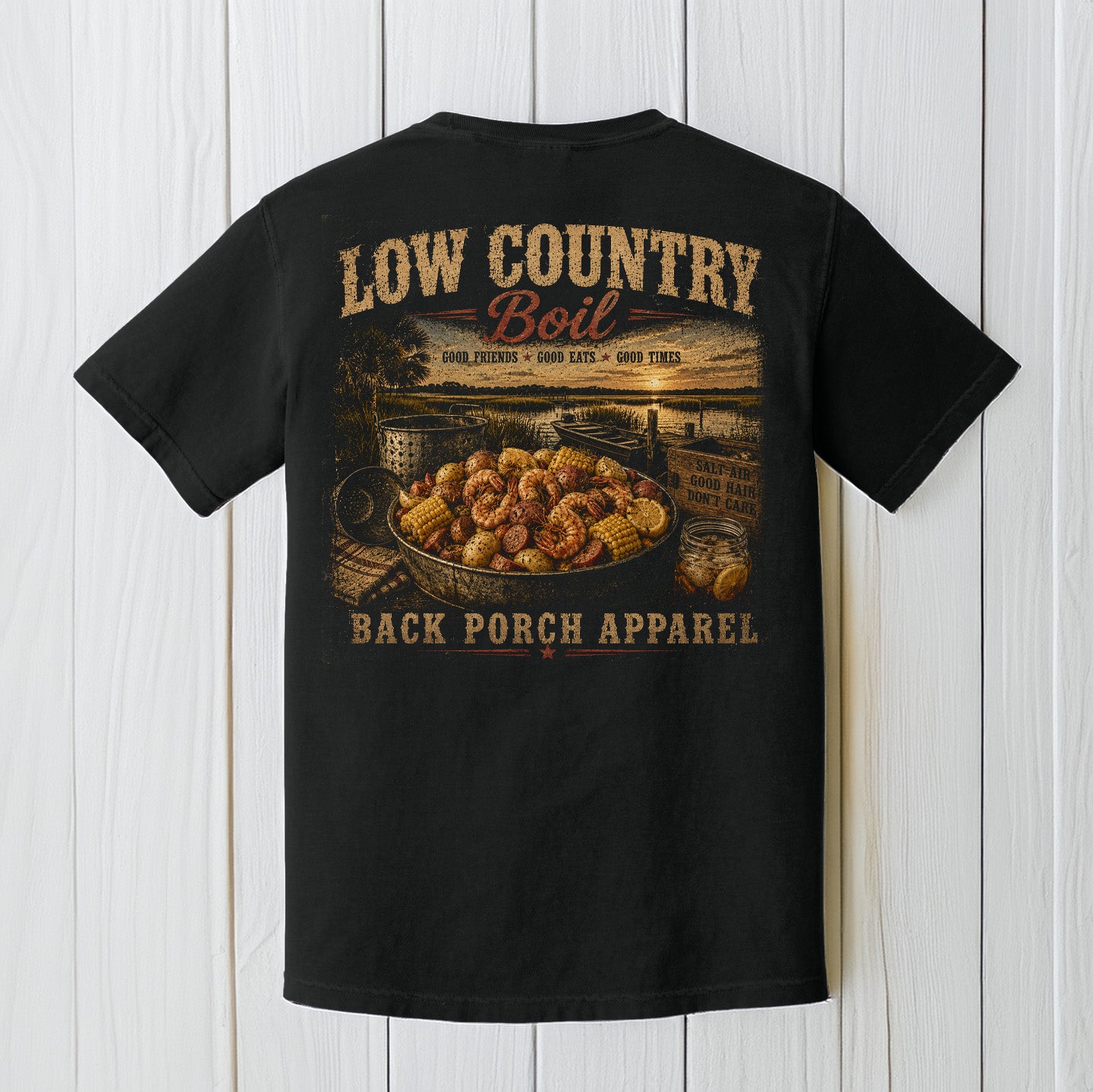 Black Comfort Colors t-shirt displayed on a wood background featuring a low country boil seafood graphic with rustic coastal southern artwork.