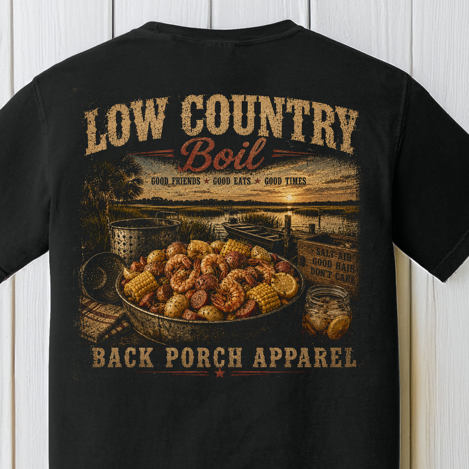 Distressed southern coastal graphic featuring a low country boil with shrimp, sausage, corn, and waterfront sunset scenery on a black Comfort Colors shirt.