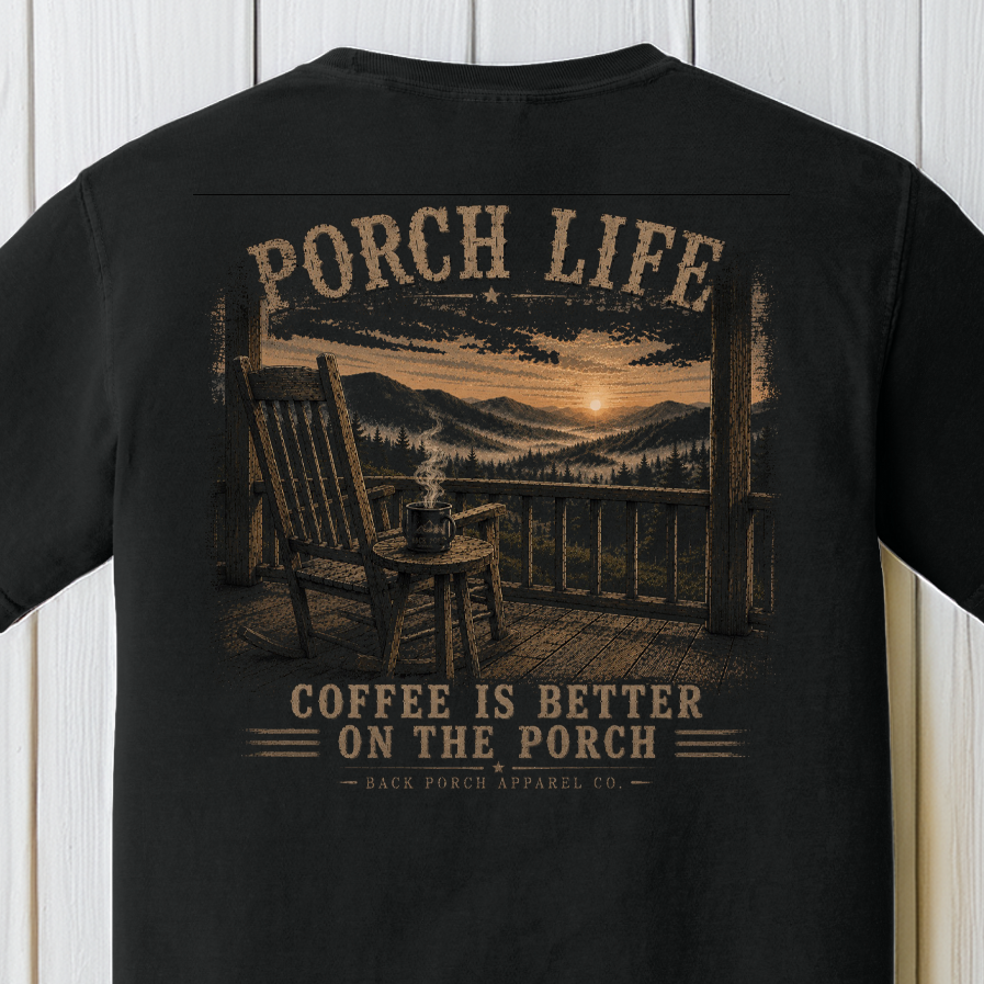 Close-up of the Porch Life Coffee Is Better on the Porch artwork showing a rocking chair, steaming coffee mug, mountain sunrise, and scenic porch view on a Black Comfort Colors t-shirt.