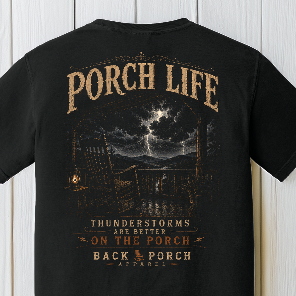 Close-up of the Porch Life Thunderstorms Are Better on the Porch artwork showing a rocking chair, lantern, mountain thunderstorm, and lightning on a Black Comfort Colors t-shirt.