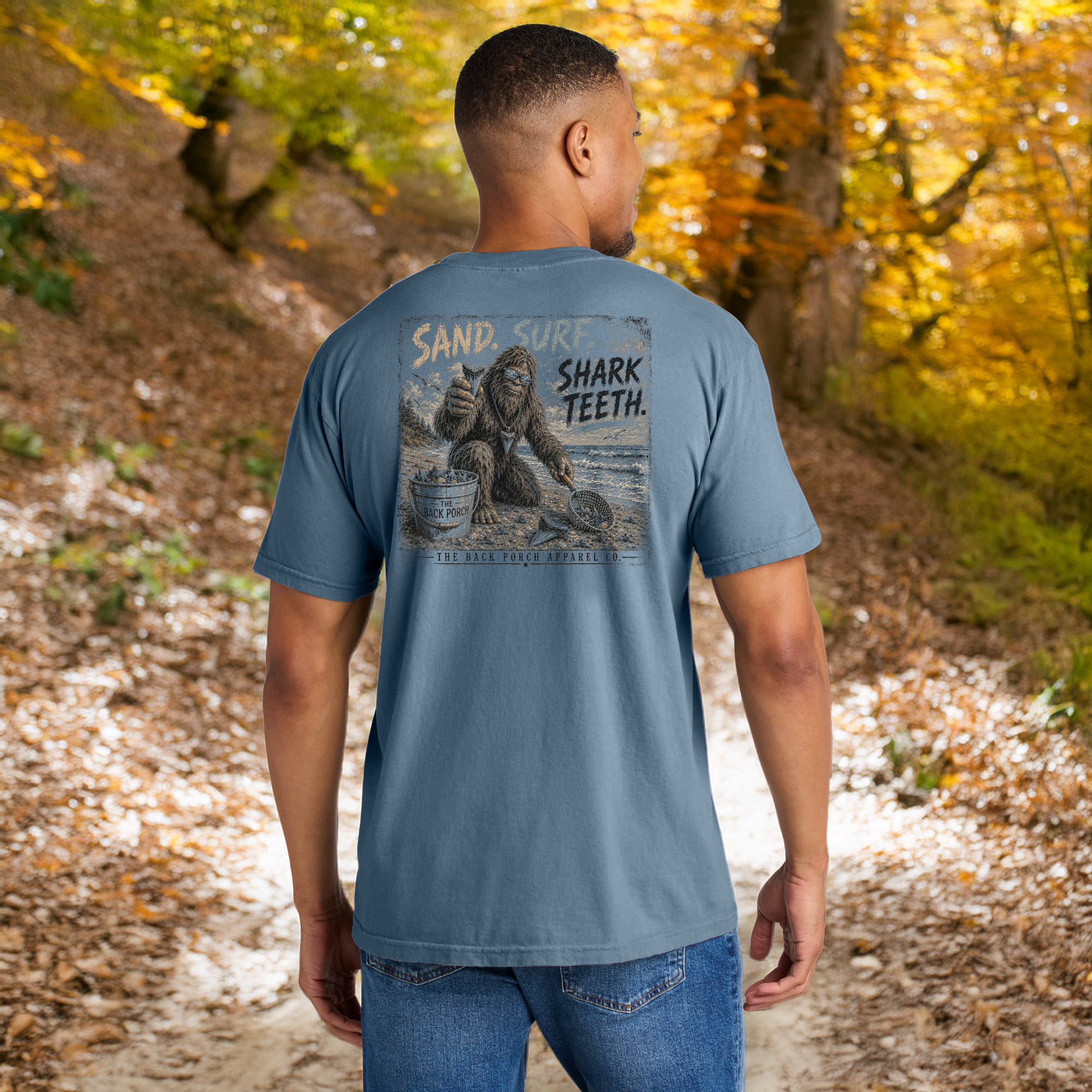 Lifestyle image of a model wearing the Back Porch Apparel Co. Sand. Surf. Shark Teeth. Comfort Colors Blue Jean graphic t-shirt featuring a large distressed Bigfoot shark tooth hunting design on the back.