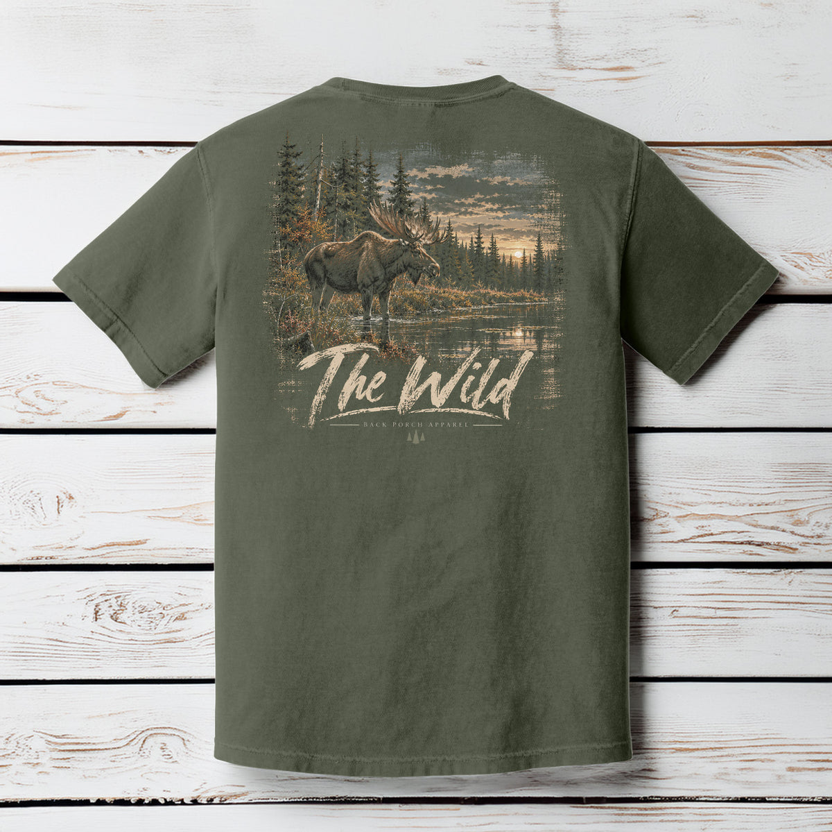 An olive green short-sleeve t-shirt shown from the back, featuring a large graphic print of a moose in a forest by a lake with the text 'The Wild' and 'Black Forest Apparel' printed below it. The shirt is displayed flat against a white-washed wooden plank background.