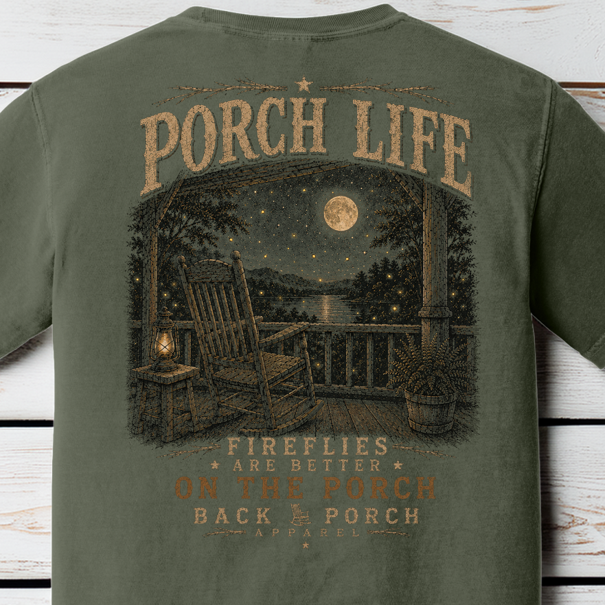 Close-up of the Porch Life Fireflies Are Better on the Porch artwork showing a rocking chair, lantern, moonlit lake, and glowing fireflies on a Moss Comfort Colors t-shirt.