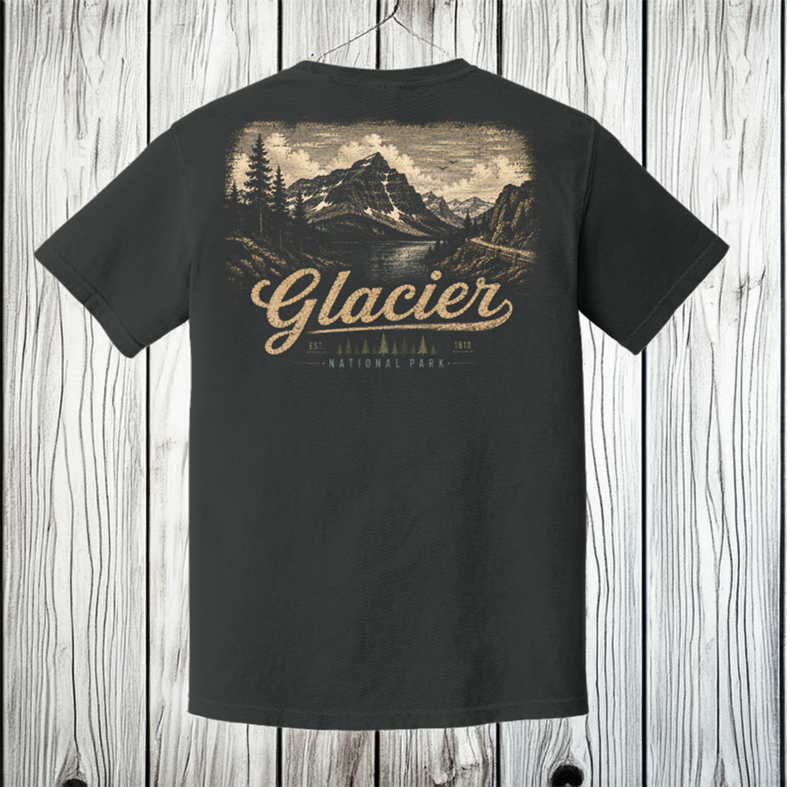 A black short-sleeve t-shirt shown from the back, featuring a large vintage-style graphic of Glacier National Park with mountains, trees, and a lake, printed in beige and brown tones. The t-shirt is hanging on a wooden hanger against a light gray wood plank background.