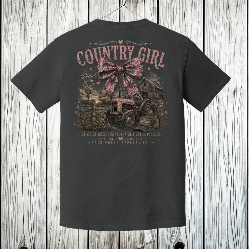Pepper Comfort Colors t-shirt hanging on a wood background featuring a large distressed country girl graphic with a pink camo bow, tractor, barn, and rustic southern design.