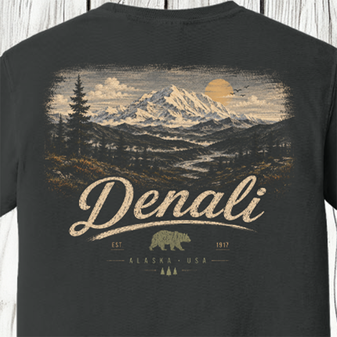 A close-up of a dark charcoal gray t-shirt showing the back side with a detailed graphic print of a mountain landscape and the word 'Denali' in cream-colored lettering.