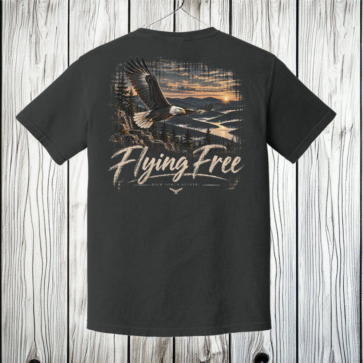 A dark charcoal gray t-shirt with a large graphic print on the back, featuring a bald eagle flying over a lake at sunset with the text 'Flying Free' and 'Mendota Wilderness'. The shirt is displayed against a rustic white-washed wood plank background.