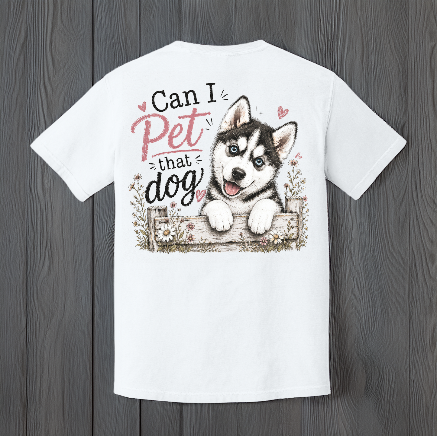 White unisex t-shirt featuring a "Can I Pet That Dog" graphic with a smiling blue-eyed husky puppy peeking over a rustic wooden fence, displayed on a wood background.