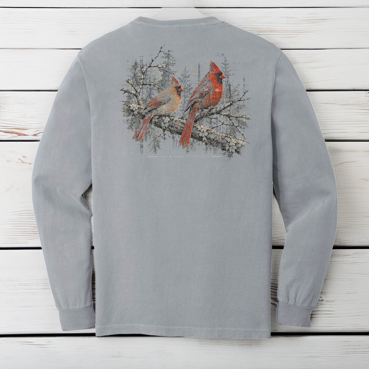 A light gray long-sleeve t-shirt shown from the back, featuring a graphic print of two red cardinals perched on a snowy branch. The shirt is laid flat on a white wooden plank background.