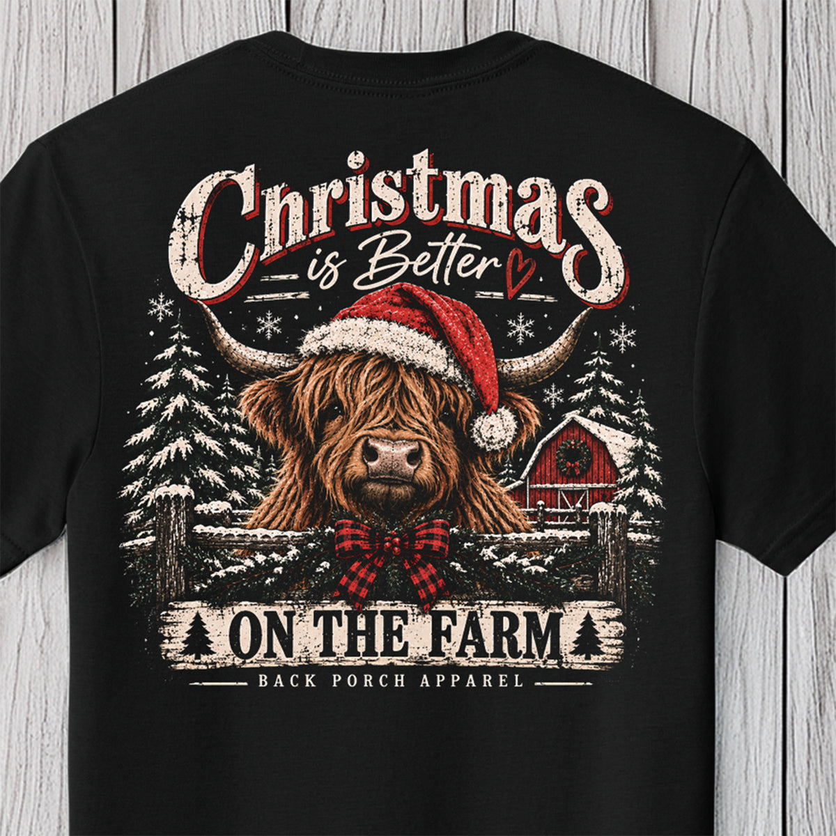 A black short-sleeve graphic t-shirt displayed flat against a light gray wood plank background. The shirt features a festive Christmas design with a highland cow wearing a red Santa hat and red plaid bow tie, surrounded by snow-covered pine trees, a red barn, and distressed text reading 'Christmas is Better ON THE FARM' with 'BACK PORCH APPAREL' printed below.