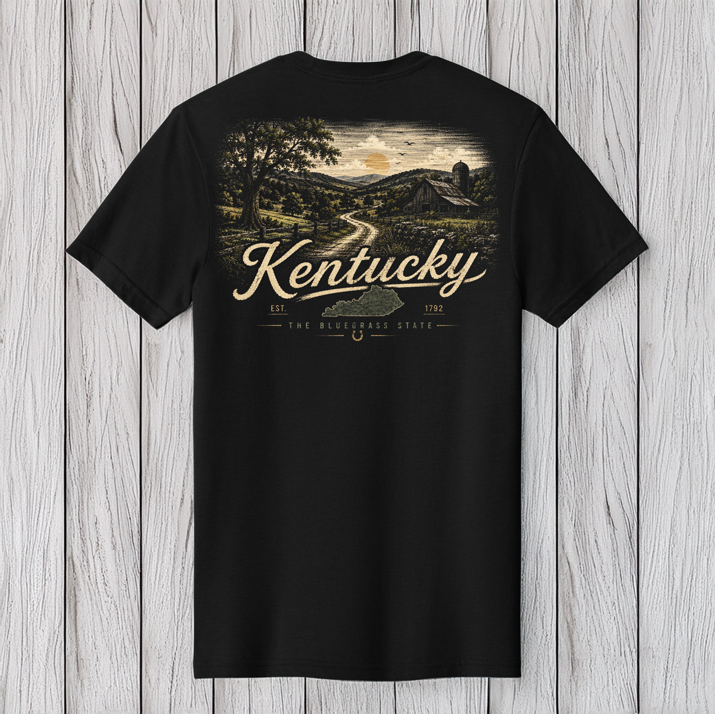A black short-sleeve t-shirt with a vintage-style Kentucky landscape graphic on the front, featuring a barn, trees, and a winding road under a sunset sky, with 'Kentucky' written in large cream script. The t-shirt is displayed flat against a light gray wood plank background.
