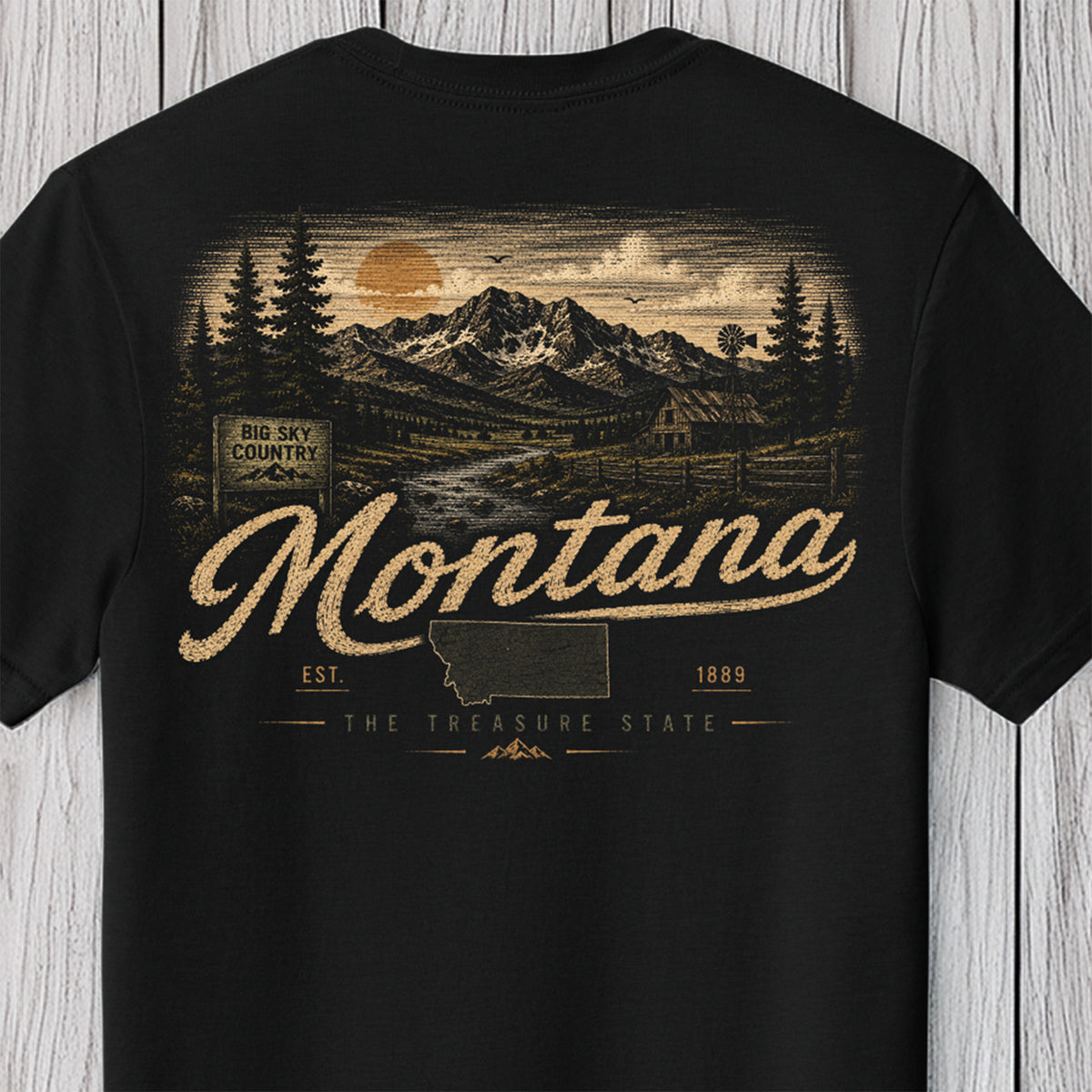 A black short-sleeve t-shirt with a vintage-style Montana graphic on the front, featuring a mountain landscape, evergreen trees, and the word 'Montana' in large script, displayed against a light gray wood plank background.