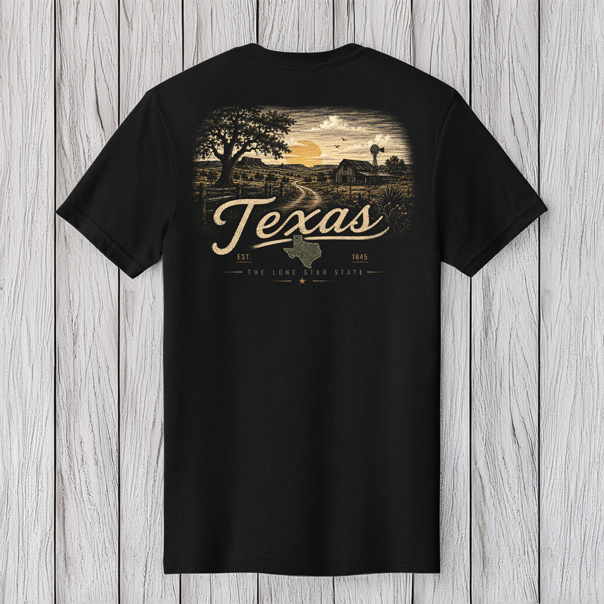 A black short-sleeve t-shirt with a vintage-style Texas landscape graphic on the front, featuring a sunset, trees, a barn, and the word 'Texas' in large script, displayed against a light gray wood plank background.