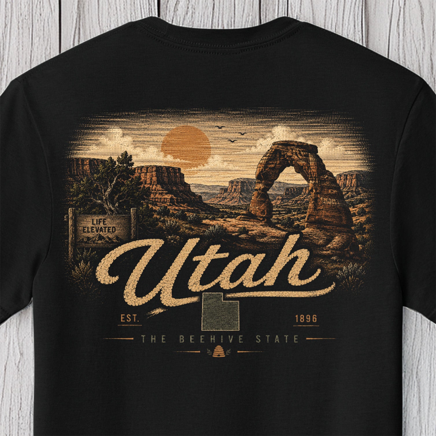 A black short-sleeve t-shirt with a detailed graphic print of Delicate Arch National Park in Utah, featuring the text 'Utah' and 'The Beehive State' in a vintage style, displayed against a light gray wood plank background.