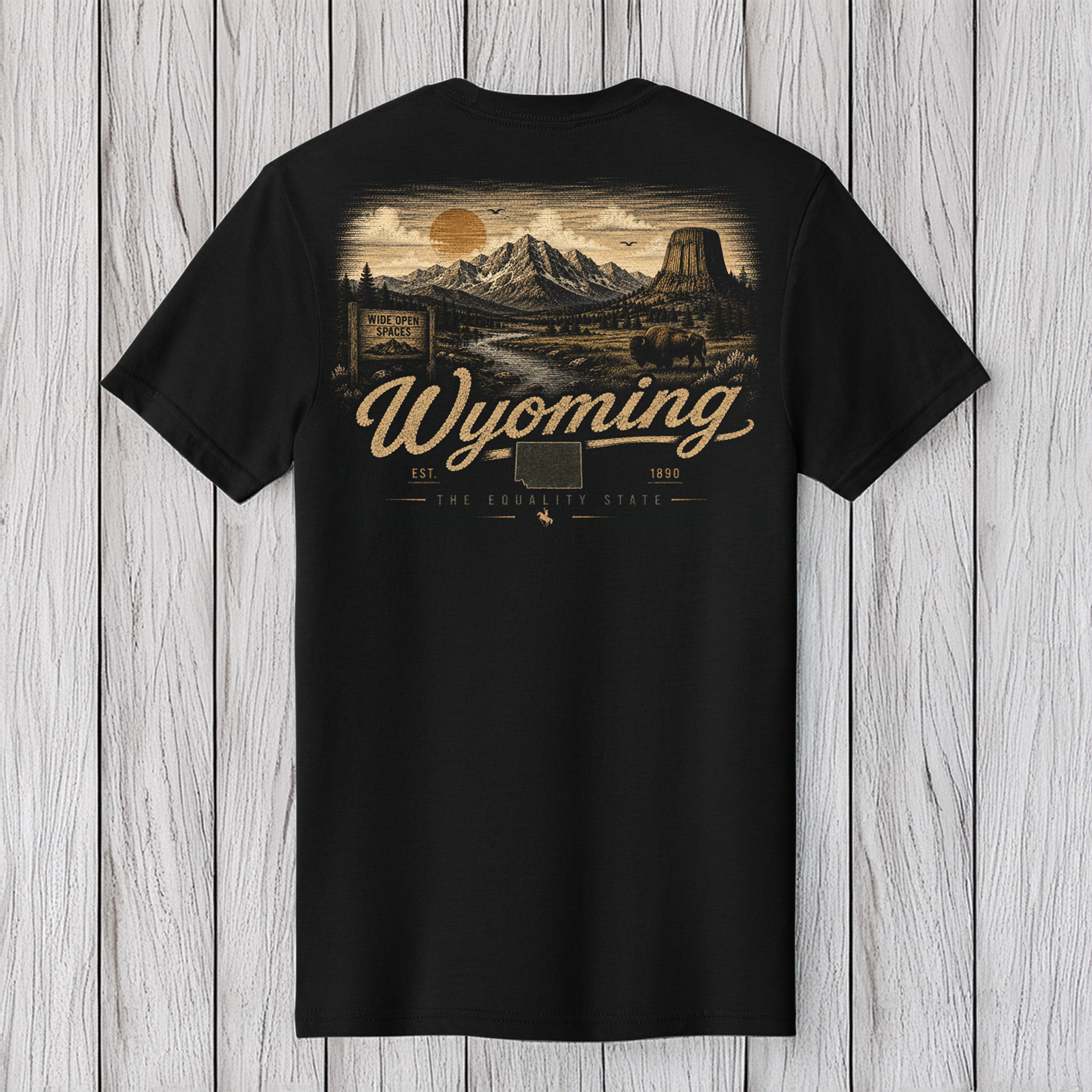 A black short-sleeve t-shirt with a detailed vintage-style Wyoming landscape graphic on the back, featuring a mountain scene, a sun, and the text 'Wyoming' in a cursive font, displayed against a light gray wood plank background.