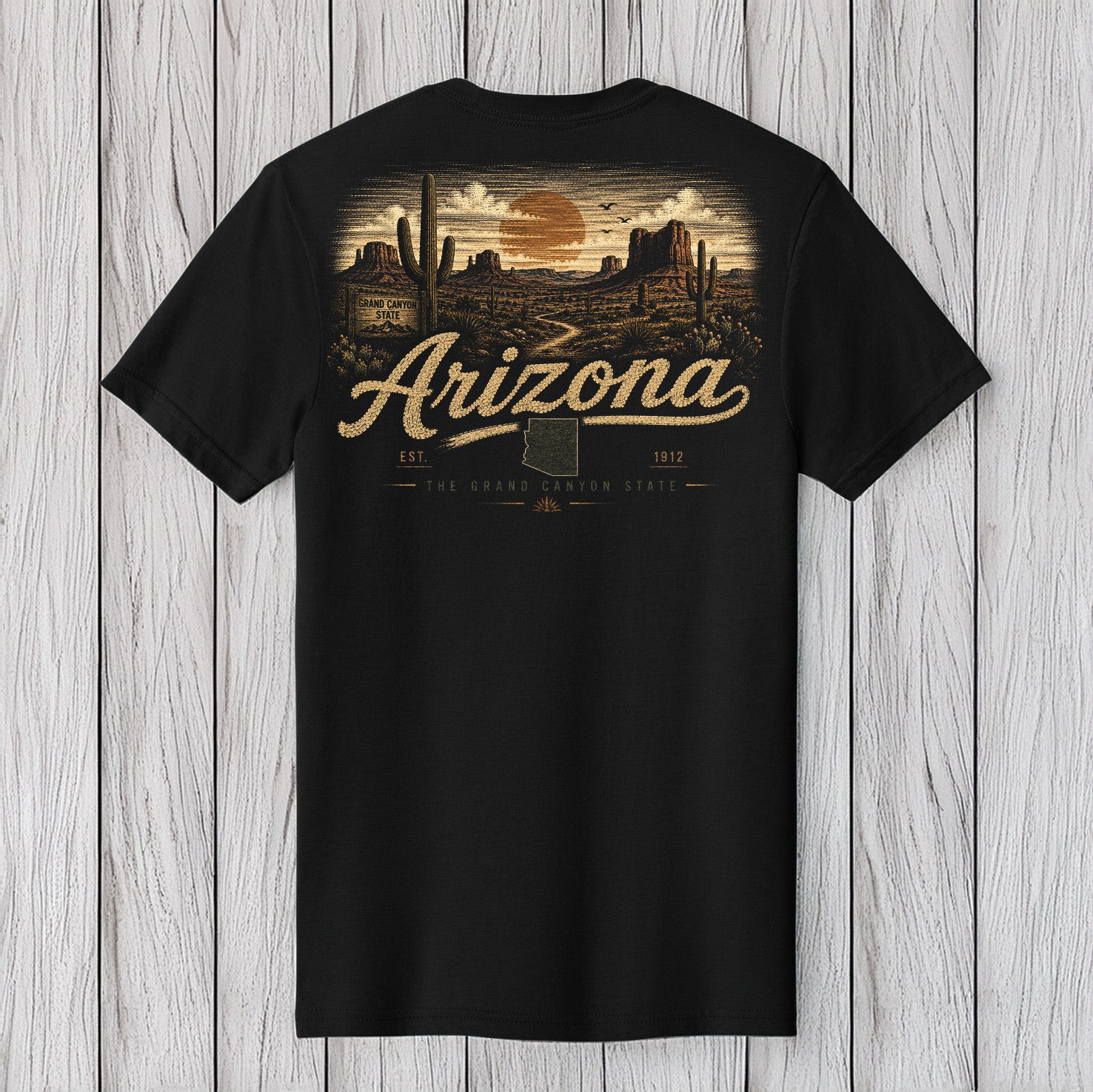 A black short-sleeve t-shirt with a detailed Arizona desert landscape graphic on the front, featuring the text 'Arizona' and 'The Grand Canyon State' in a vintage style, displayed flat against a light gray wood plank background.