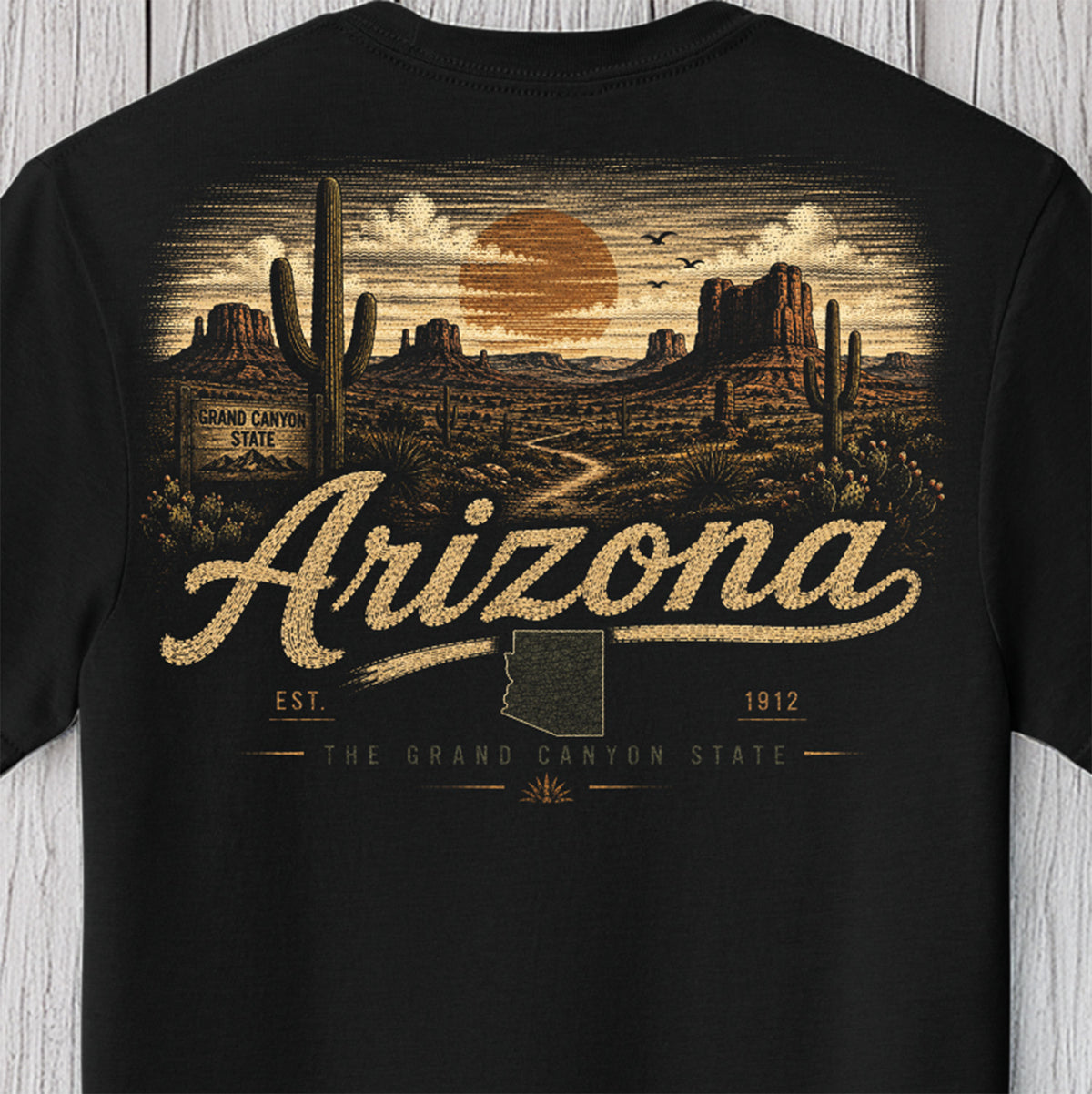 A black short-sleeve t-shirt featuring a detailed desert landscape graphic on the front. The design depicts Grand Canyon rock formations, saguaro cacti, a winding path, and a large orange sun in a cloudy sky, with 'Arizona' written in a textured beige script across the center.