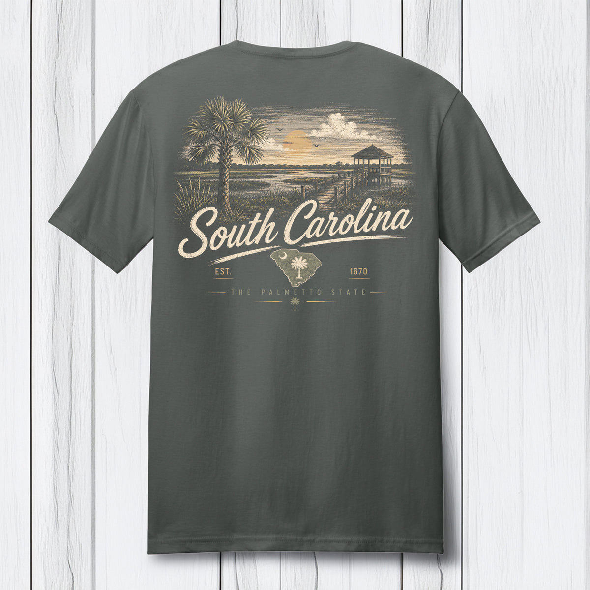 A dark gray t-shirt shown from the back, featuring a vintage-style South Carolina graphic with a palm tree, beach scene, and text, displayed against a white wood plank background.