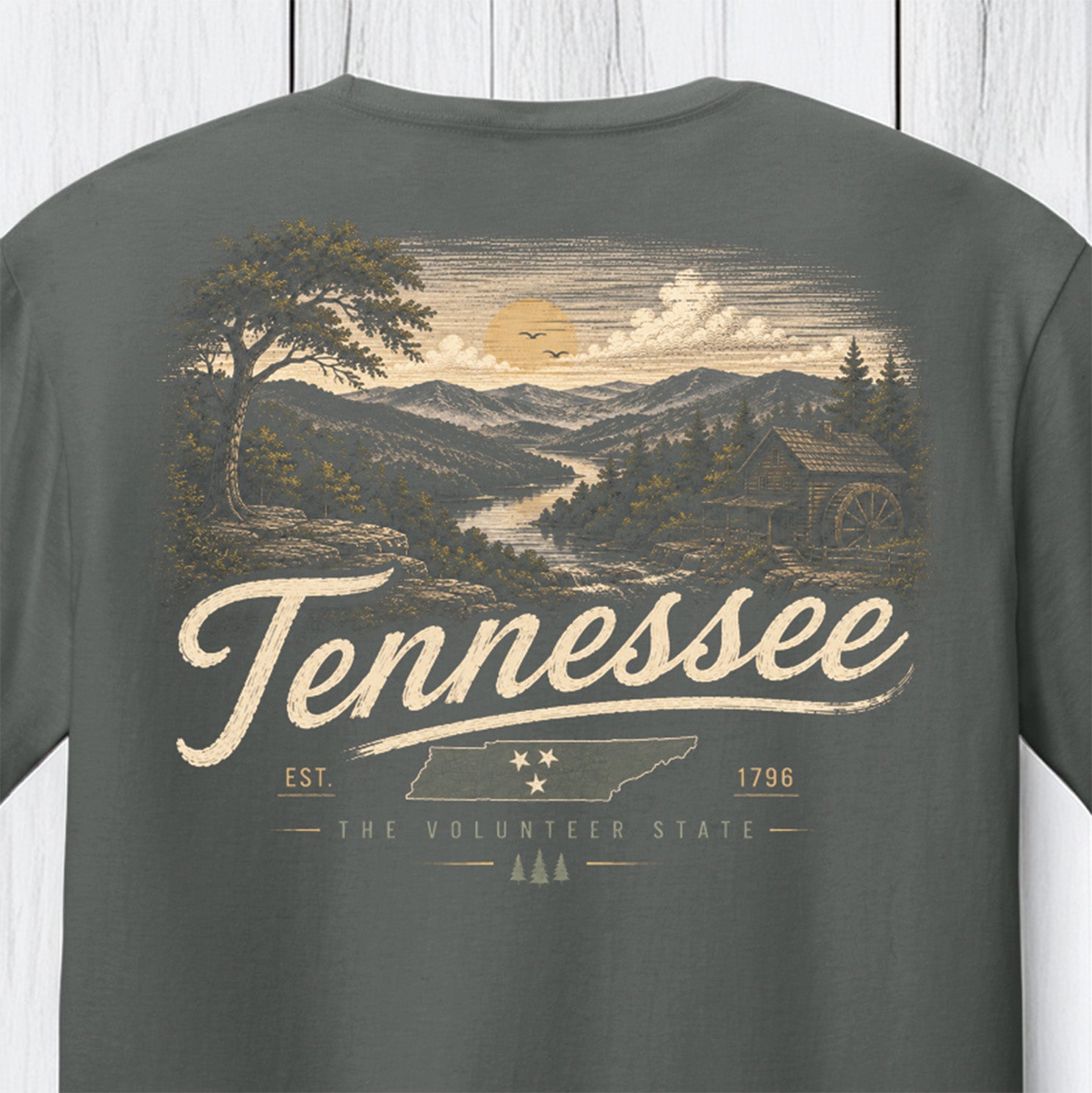 A dark gray t-shirt featuring a vintage-style Tennessee landscape graphic on the front, with a watercolor-style illustration of a mountain valley, a waterwheel, and the word 'Tennessee' in large script.