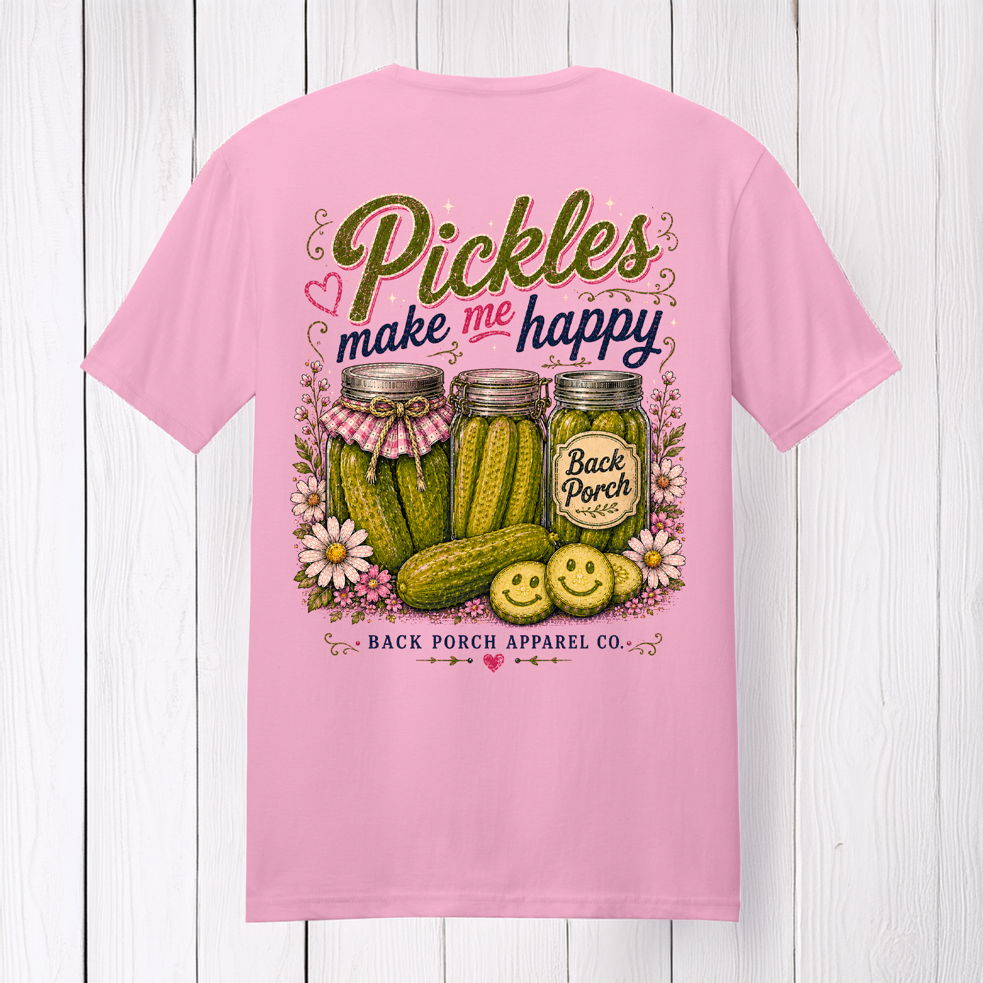 Pink 100% ring-spun cotton t-shirt featuring the "Pickles Make Me Happy" graphic with rustic pickle jars, smiling pickle slices, flowers, and Back Porch Apparel branding displayed on a white wood background.