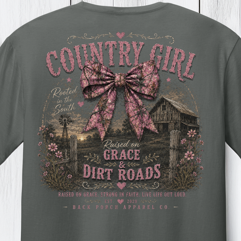 Distressed southern country girl graphic featuring a pink camouflage bow, rustic barn, wildflowers, and “Raised on Grace & Dirt Roads” typography.