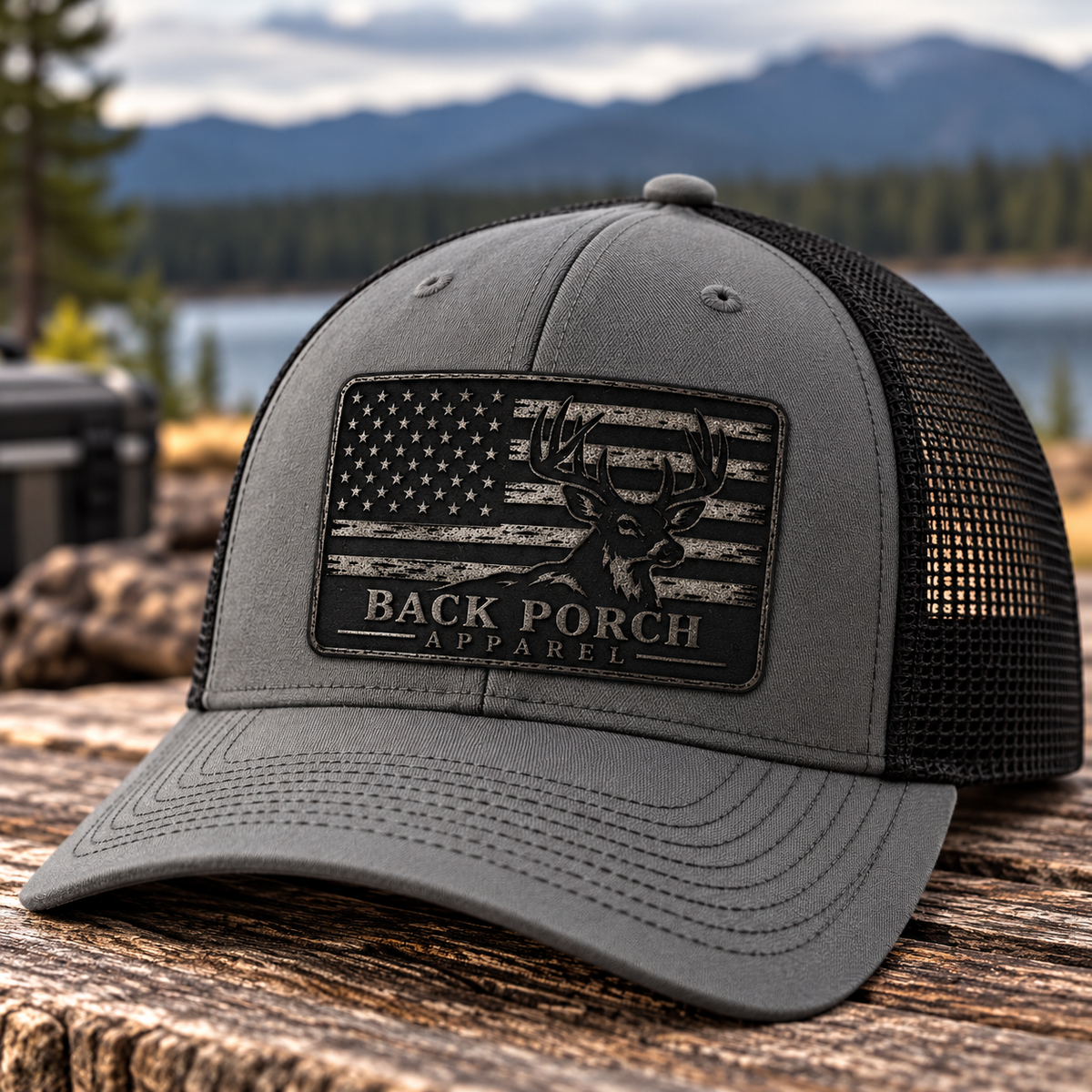 Gray and black Richardson trucker hat with charcoal leather patch featuring a whitetail buck, distressed American flag, and Back Porch Apparel logo, photographed outdoors.