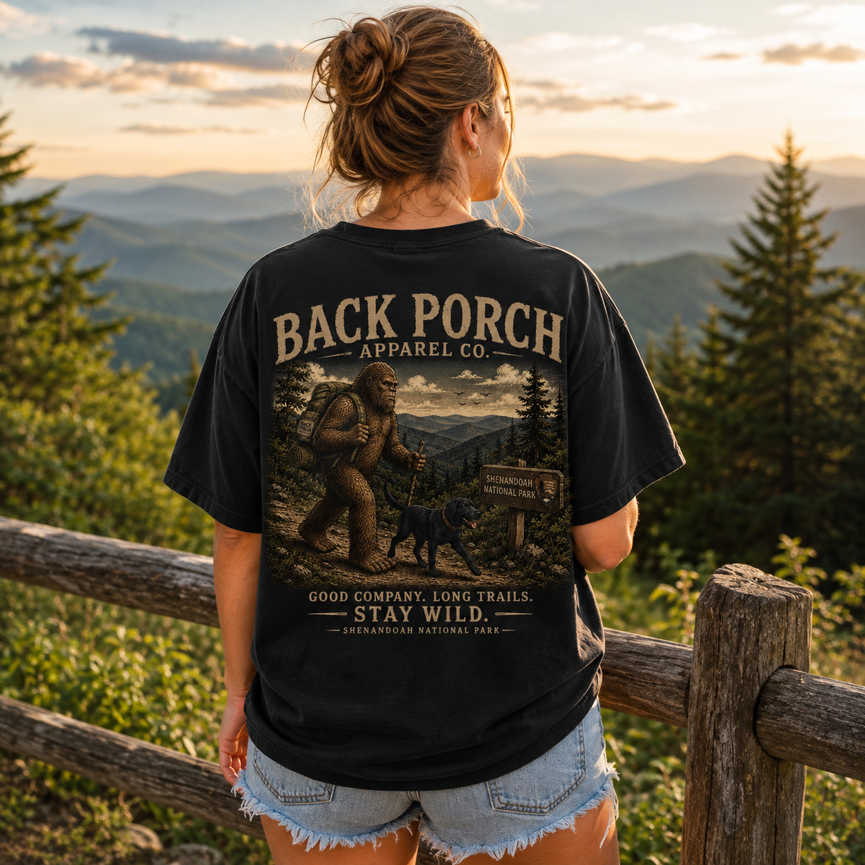 A woman stands outdoors on a mountain trail at sunset wearing a black graphic T-shirt from the back. The shirt features a large vintage-style print with the text 'BACK PORCH APPAREL CO.