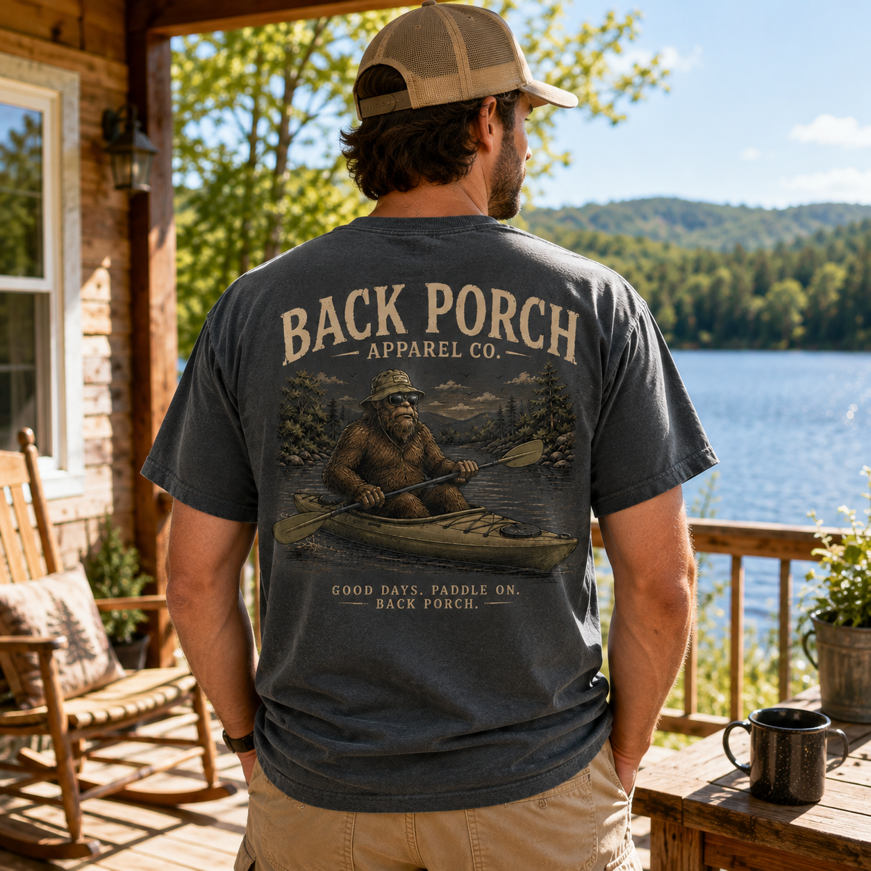 A man stands on a wooden porch by a lake, wearing a dark charcoal gray t-shirt with a large graphic print on the back. The graphic features a bigfoot in a canoe with the text 'BACK PORCH APPAREL CO.