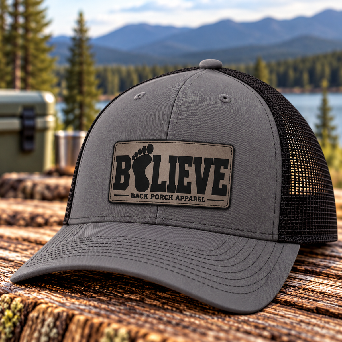 Richardson 112FP charcoal gray trucker hat with black mesh back and charcoal BELIEVE Bigfoot footprint leather patch by Back Porch Apparel, displayed outdoors beside a mountain lake.