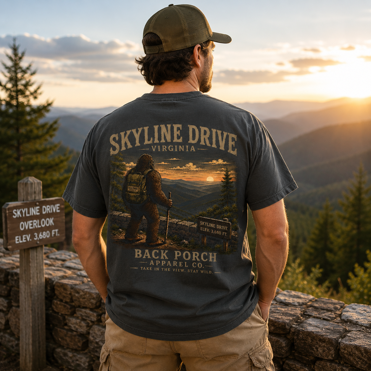 A man stands on a mountain overlook at sunset wearing a dark gray graphic t-shirt with 'SKYLINE DRIVE VIRGINIA' and a large outdoor scene printed on the back, along with khaki pants and a green cap. A wooden sign reading 'SKYLINE DRIVE OVERLOOK' is visible to the left.