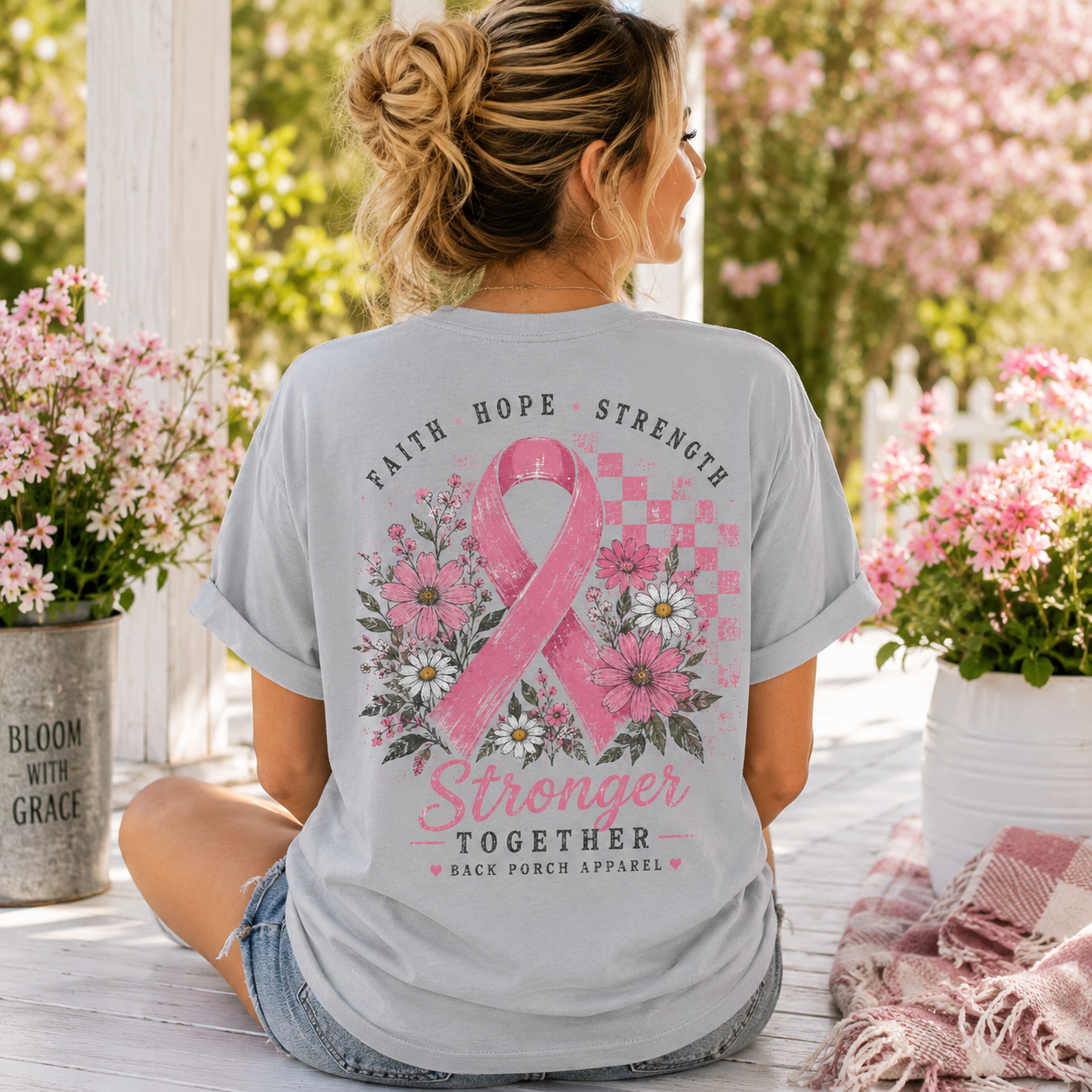 A woman sitting outdoors on a porch wearing a light gray t-shirt with a pink breast cancer awareness graphic on the back. The graphic features a pink ribbon, daisies, and the words 'FAITH HOPE STRENGTH Stronger TOGETHER BACK PORCH APPAREL'.