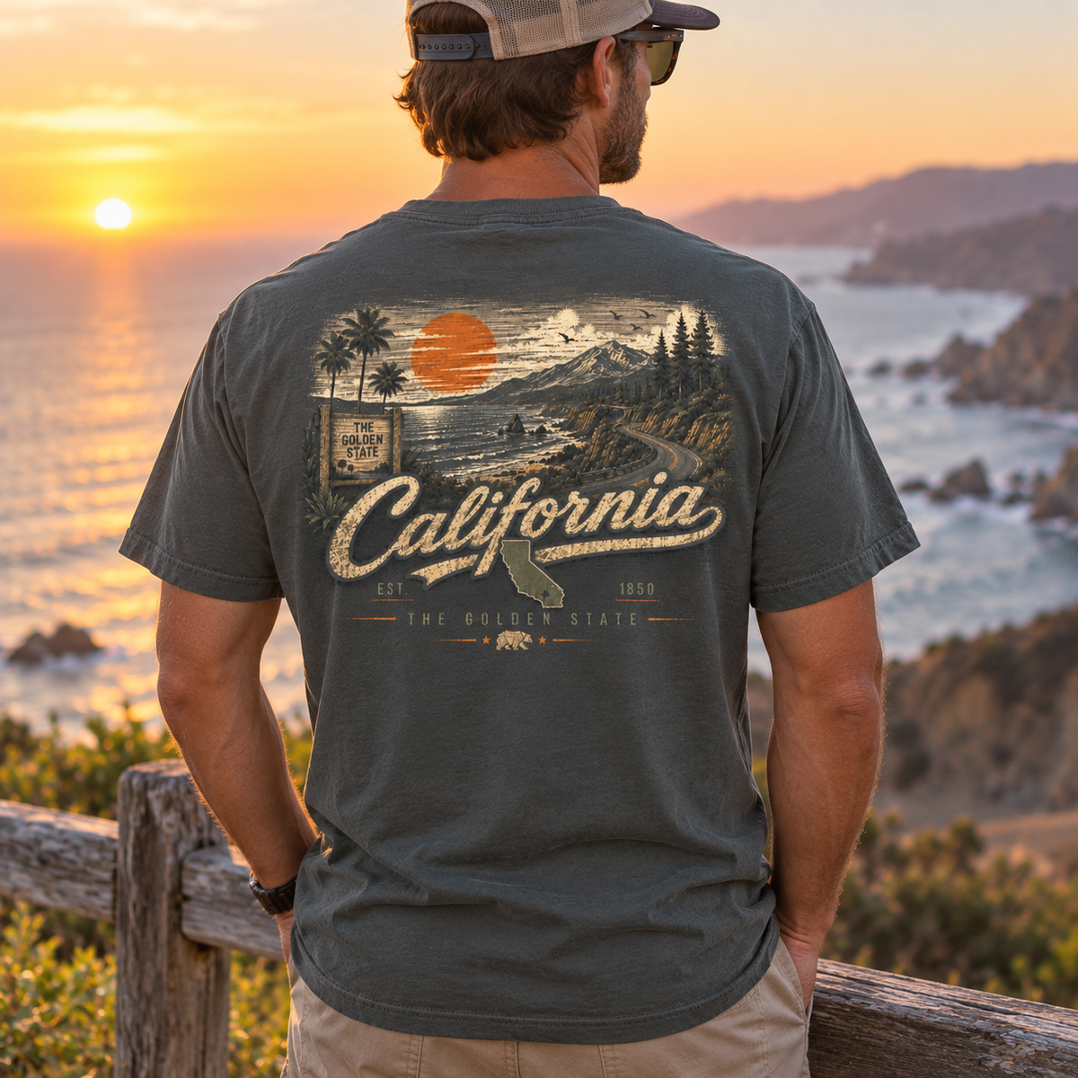 A man stands outdoors at sunset wearing a dark charcoal gray t-shirt with a large vintage-style California graphic on the back, overlooking a coastal landscape.