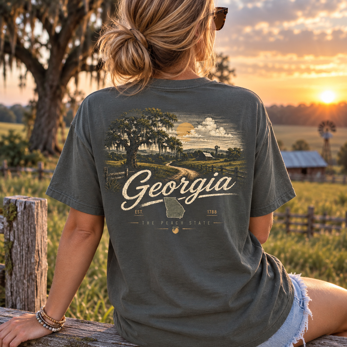 A woman sitting outdoors at sunset wearing a dark olive green graphic T-shirt with a large back print reading Georgia and showing a rural landscape scene. The shirt is the product being modeled in a real outdoor environment, so this is Lifestyle rather than Product/Packaging.