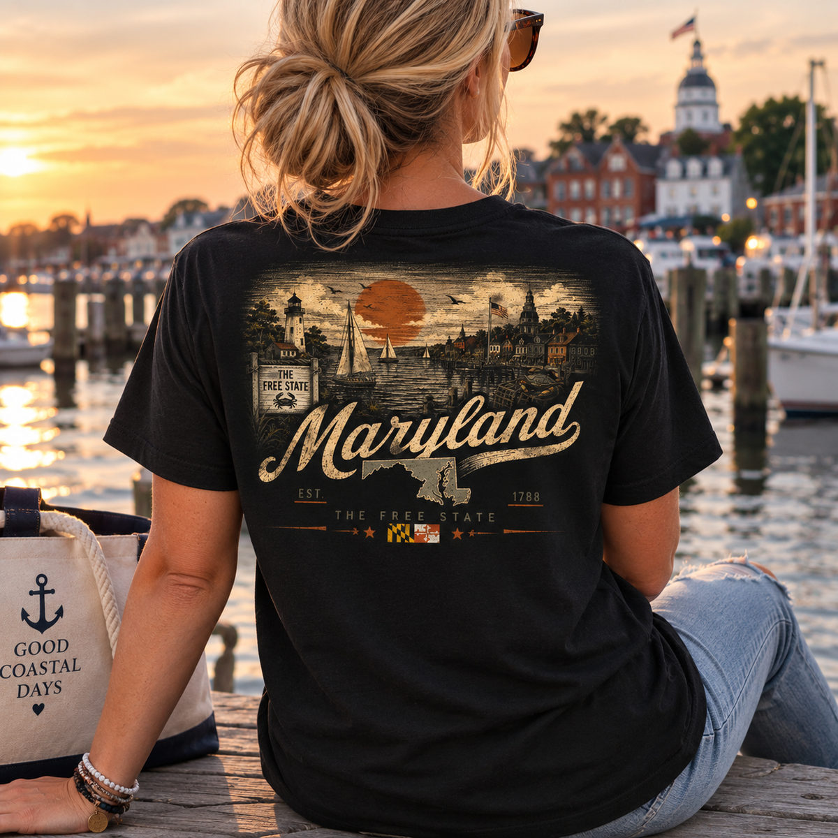 A woman sits on a dock at sunset wearing a black graphic T-shirt with a vintage-style Maryland print on the back. The shirt features an orange sun, lighthouse, boats, and the words 'Maryland' and 'The Free State.