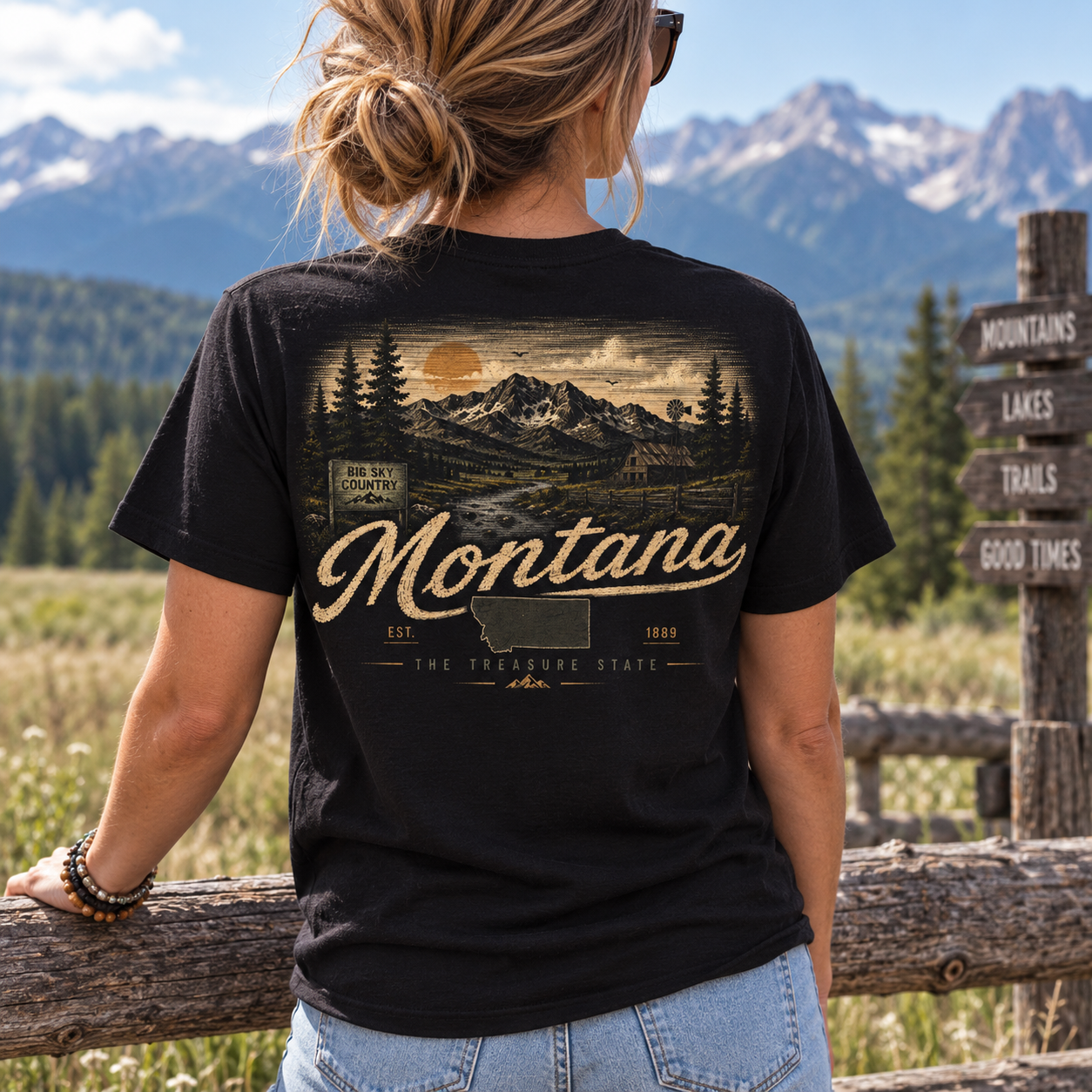 A woman wearing a black graphic t-shirt with a vintage-style Montana mountain landscape print, photographed from behind in an outdoor mountain setting.
