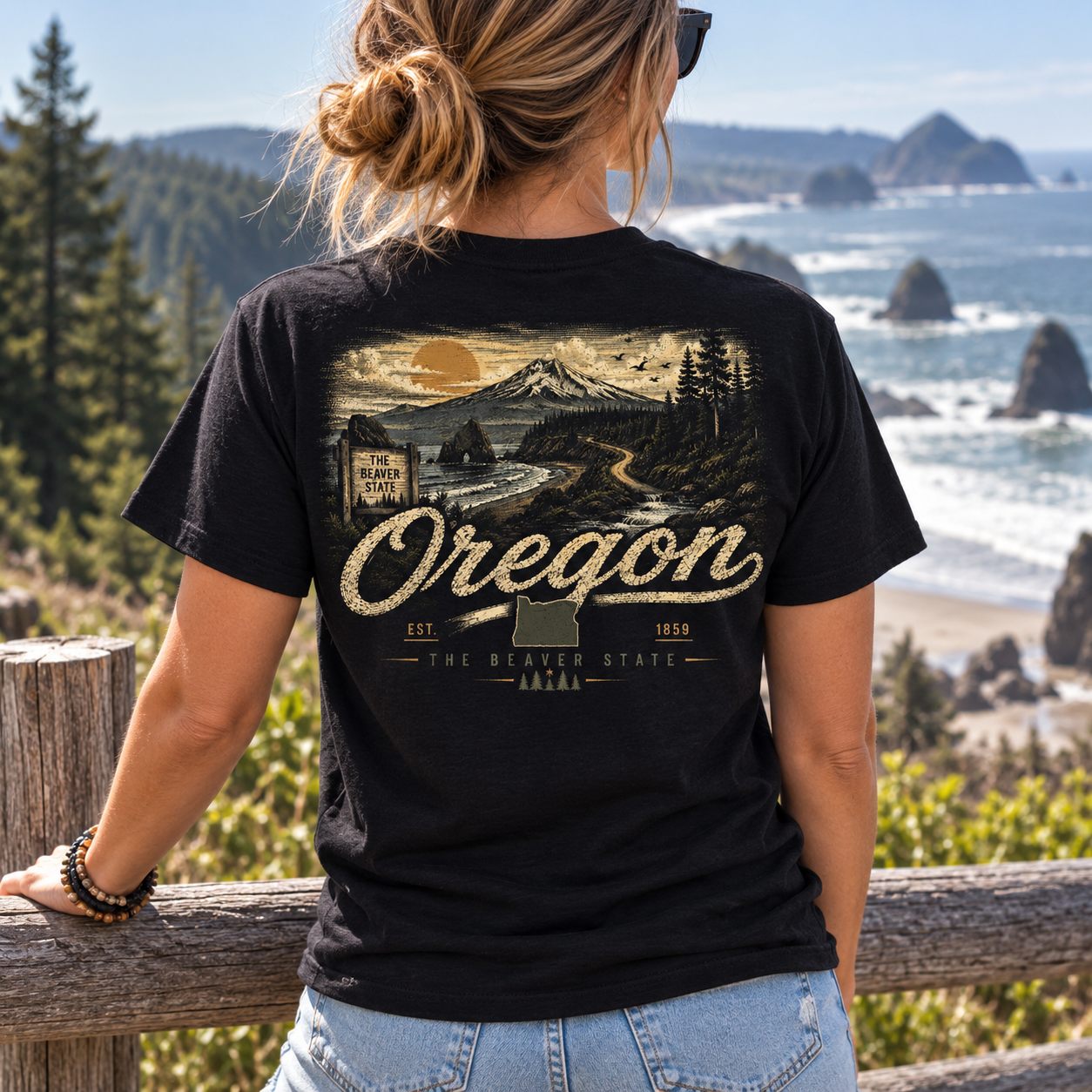 A woman viewed from behind wearing a black graphic t-shirt with a vintage-style Oregon landscape print, standing on a wooden railing with a scenic ocean coastline in the background.