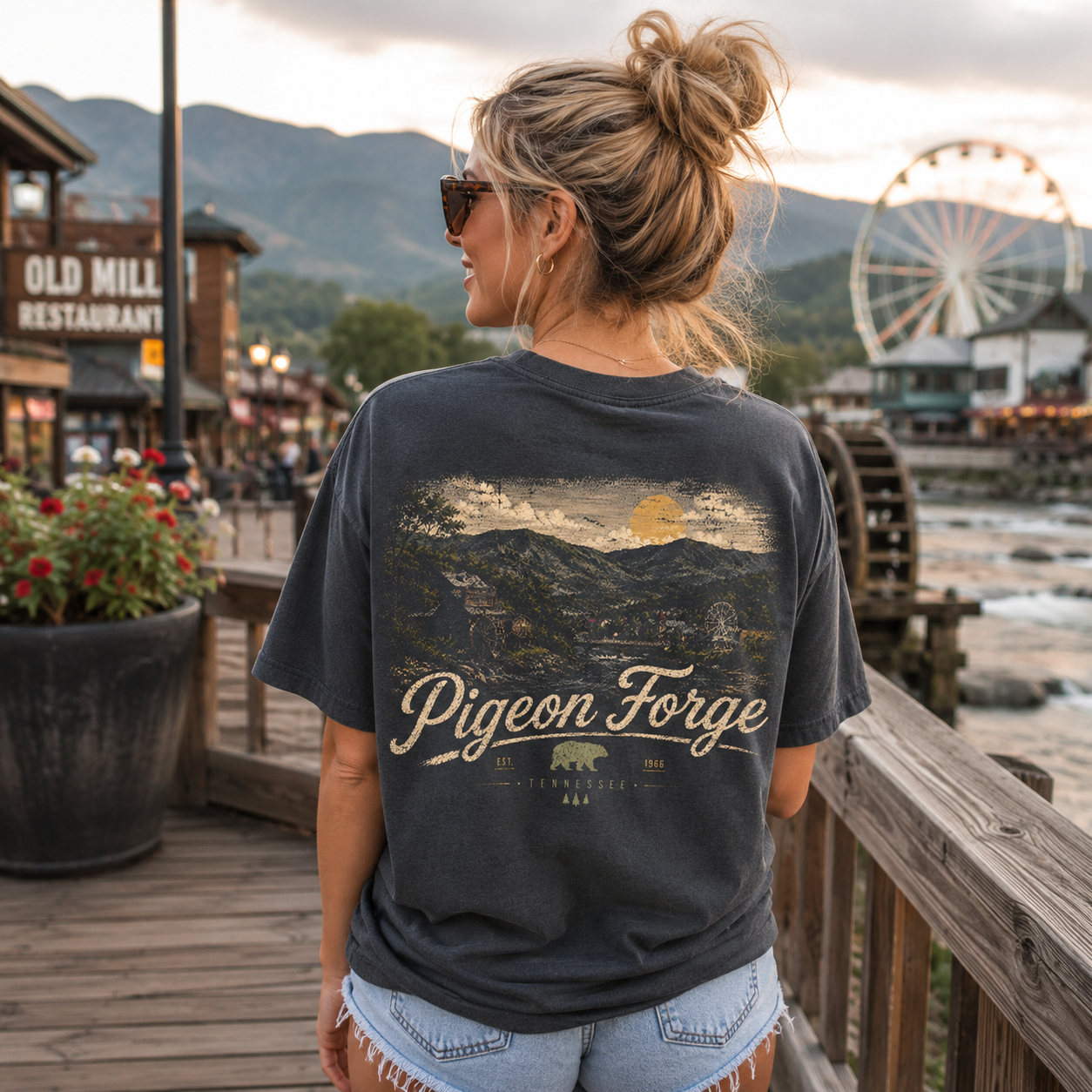 A woman stands on a wooden boardwalk wearing a dark charcoal graphic t-shirt with a mountain landscape print and the words 'Pigeon Forge' on the back. The background shows a scenic outdoor setting with a Ferris wheel, water, and buildings at sunset.