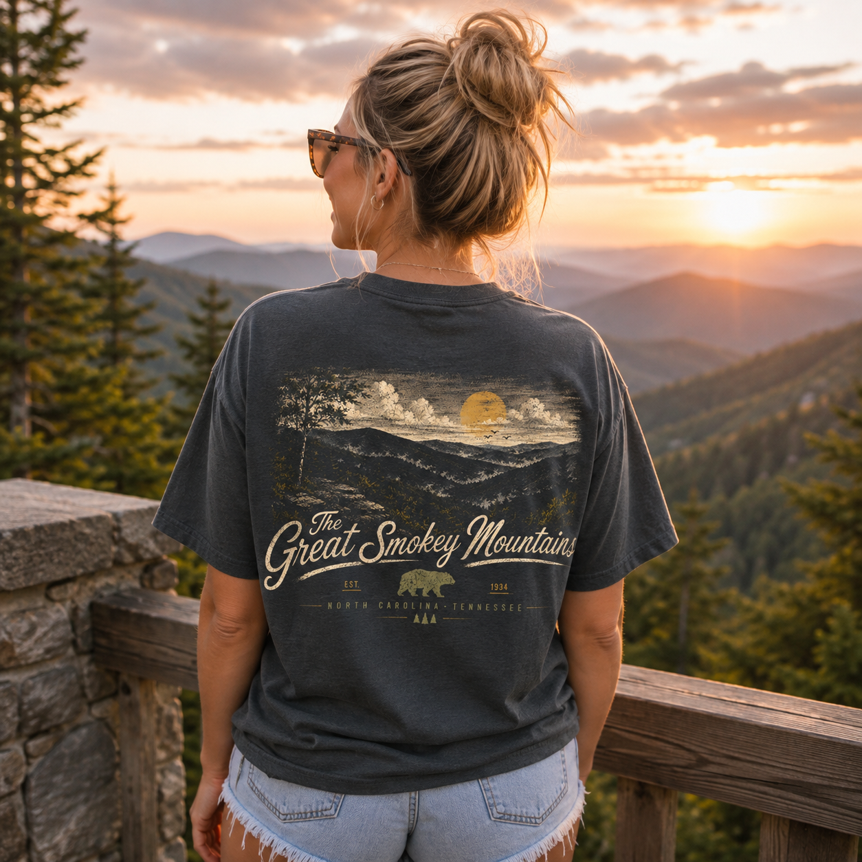 A woman stands on a wooden railing overlooking a mountain landscape at sunset, wearing a dark charcoal gray t-shirt with a large graphic print on the back that reads 'The Great Smokey Mountains' with a bear and mountain scene. She is wearing denim shorts.
