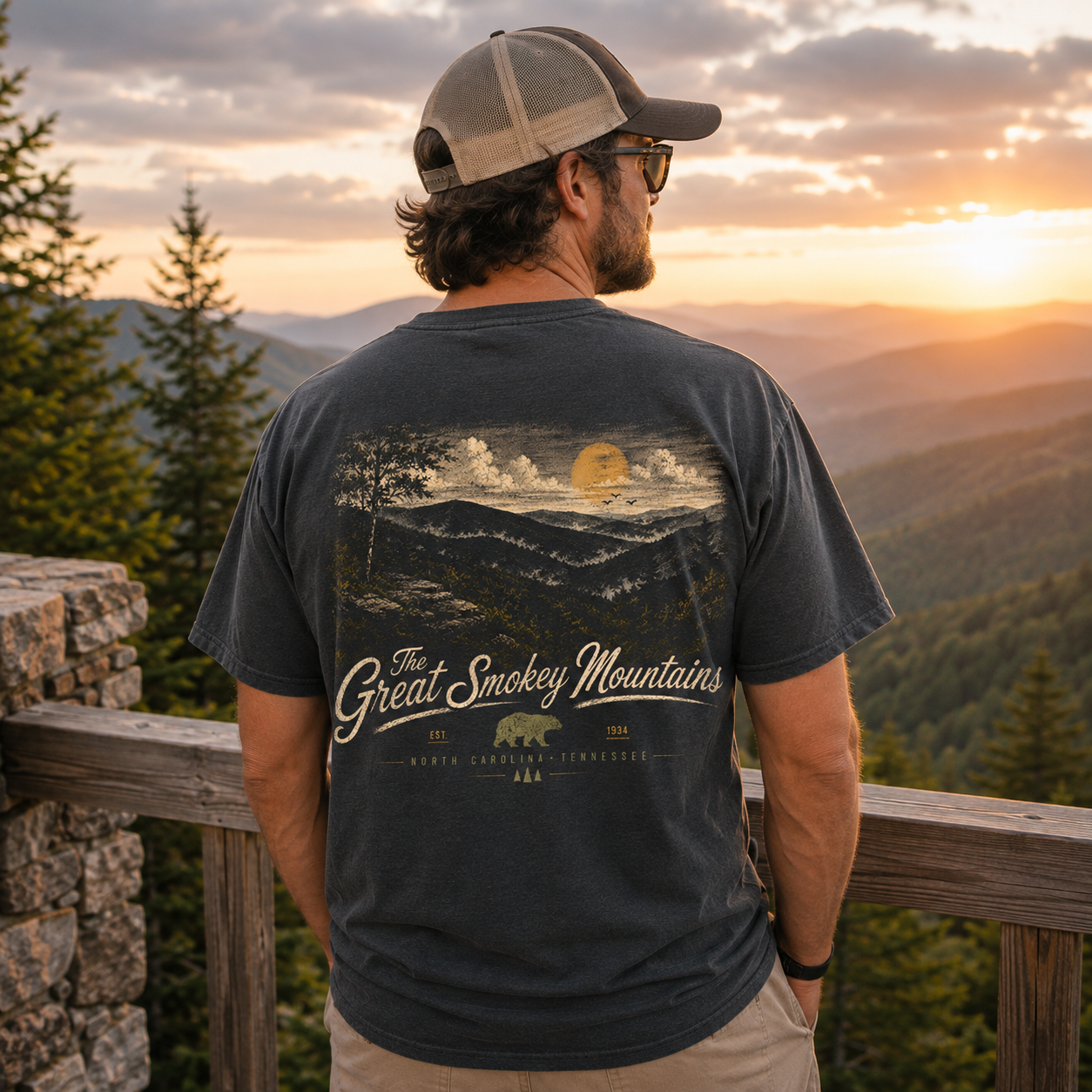 A man wearing a dark charcoal gray t-shirt with a large graphic print of 'The Great Smokey Mountains' is seen from behind, standing on a wooden railing overlooking a mountain landscape at sunset.