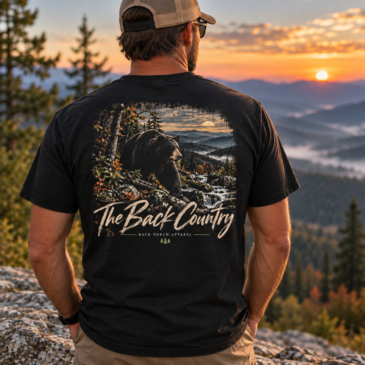 A man wearing a black graphic t-shirt with a bear and mountain landscape print on the back, standing on a rocky mountain peak at sunset. The t-shirt features the text 'The Back Country' and 'BACK DOOR APPAREL'.