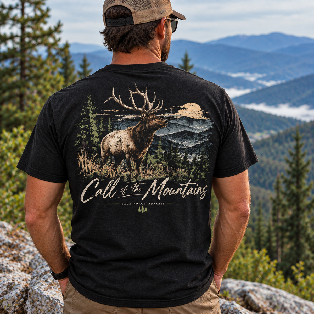 A man viewed from behind wearing a black graphic t-shirt with a large back print of an elk in a mountain landscape and the text 'Call of the Mountains'. He is outdoors on a rocky mountain peak with pine trees and blue mountains in the background.