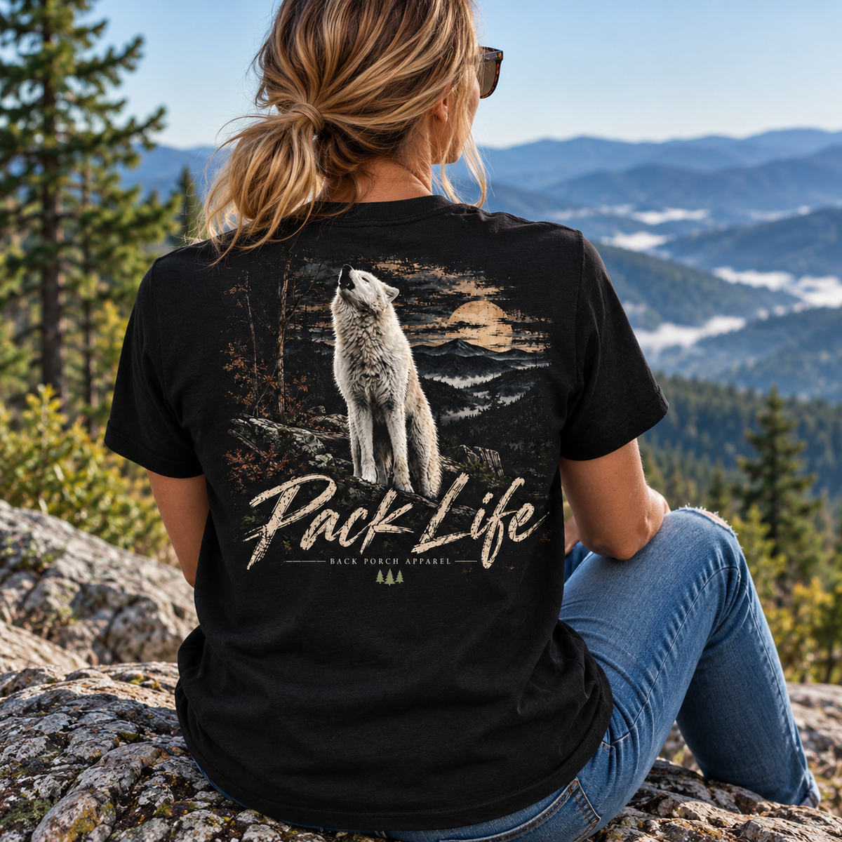 A woman wearing a black graphic t-shirt with a large back print of a howling wolf and the text 'Pack Life', photographed from behind against a mountain landscape.