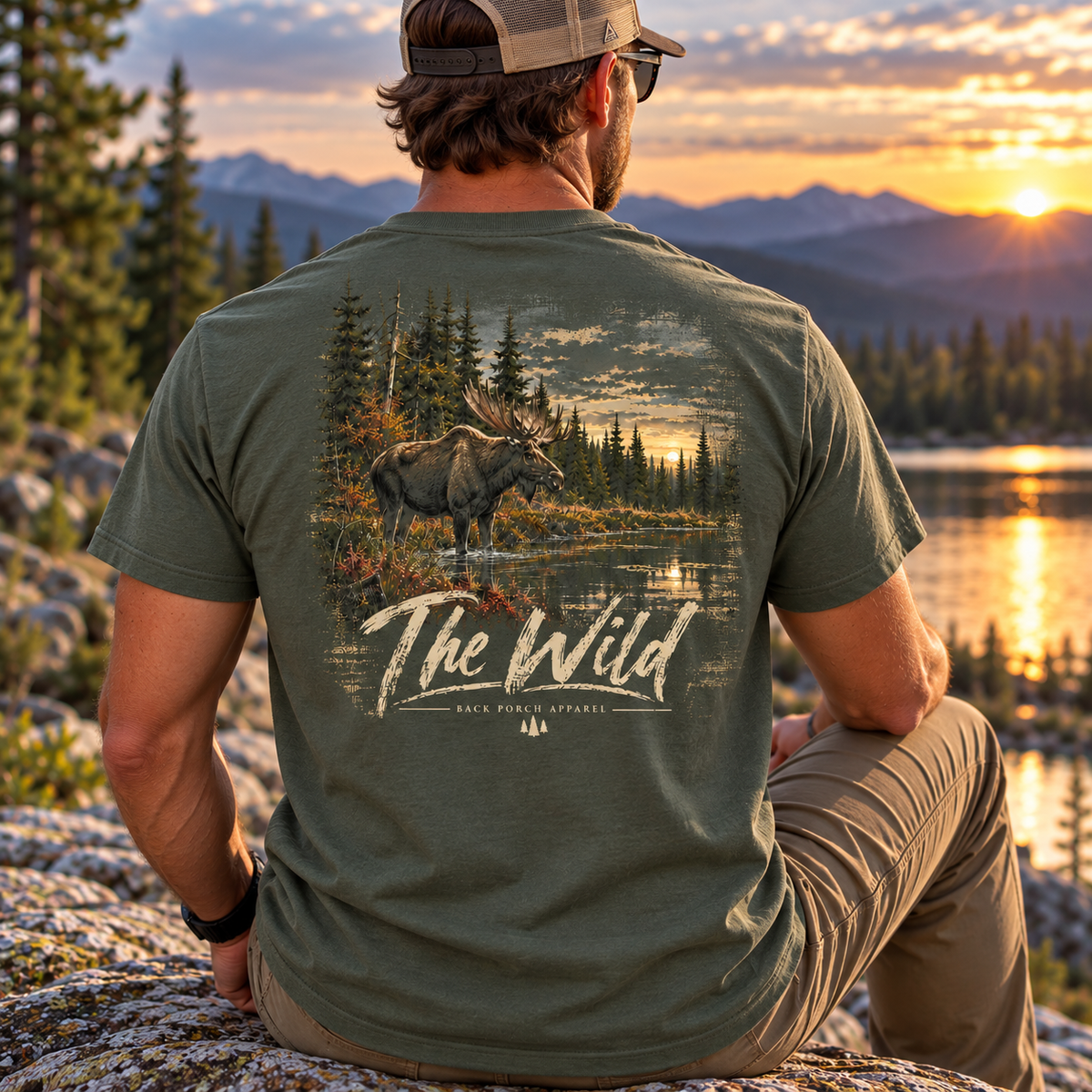 A man sits outdoors on a rock at sunset facing a mountain lake landscape, wearing an olive green graphic T-shirt with a large back print of a moose in a forest and the words “The Wild.”.